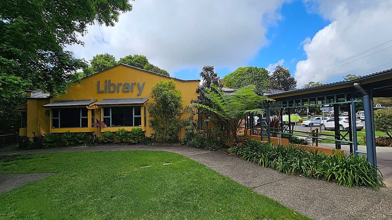A yellow building with the word "Library" on the front is on the left, surrounded by lush greenery and plants. A pathway leads to the entrance, bordered by grass. On the right, there's a covered walkway with a green metal frame, leading to a car park with several vehicles visible. Trees and bushes are in the background, adding to the verdant setting.