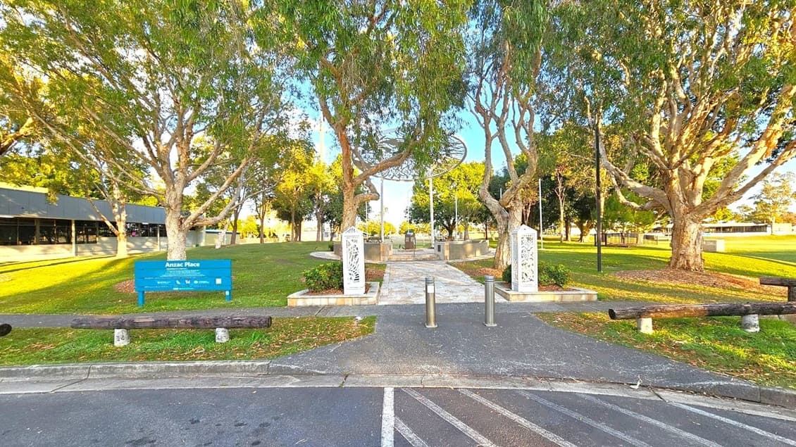 A circular stone pathway leads to a central monument with a decorative metal canopy above, featuring intricate cut-out designs. The area is surrounded by a low stone wall and benches. Trees with lush green foliage line the background, and a building with a sloped roof is visible to the left. The ground is covered with grass, and there are scattered leaves on the path.