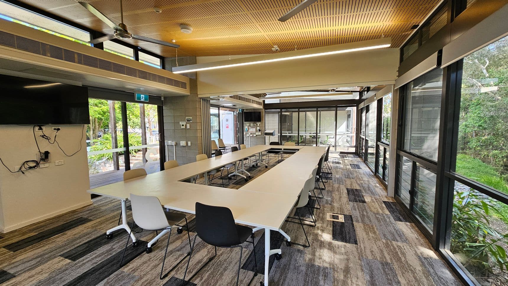 A modern conference room with a U-shaped arrangement of white tables and assorted chairs. The left side features a wall-mounted TV with visible cables. Large windows on the right and at the back provide views of greenery outside. The ceiling is wooden with recessed lighting and ceiling fans. The floor is covered with patterned carpet tiles in shades of grey and beige.