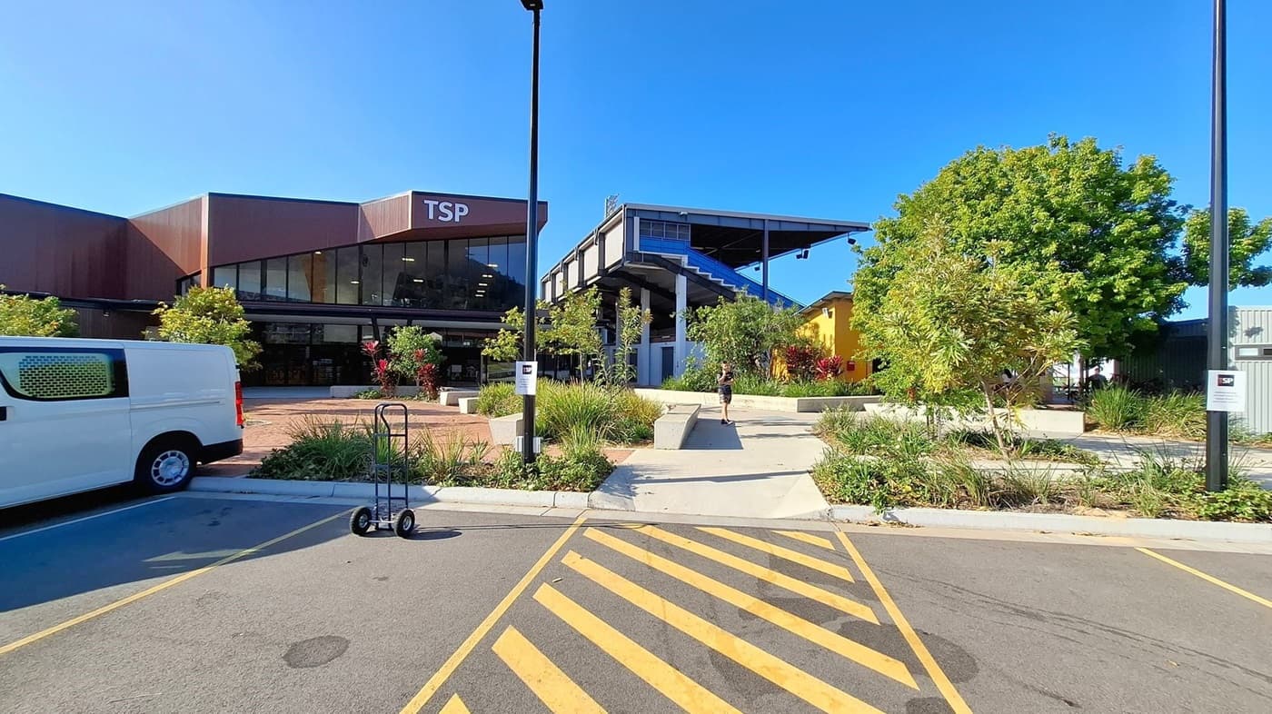 A courtyard with a central raised garden bed filled with small trees and shrubs. A person stands near the garden, casting a shadow on the ground. To the right, a building with large windows and the letters "TSP" on the facade. In the background, a car park with several vehicles and a sign reading "Townsville Sports Precinct". Overhead, the sun is bright, and tree branches with green leaves frame the top of the image. The ground is paved with a textured surface.