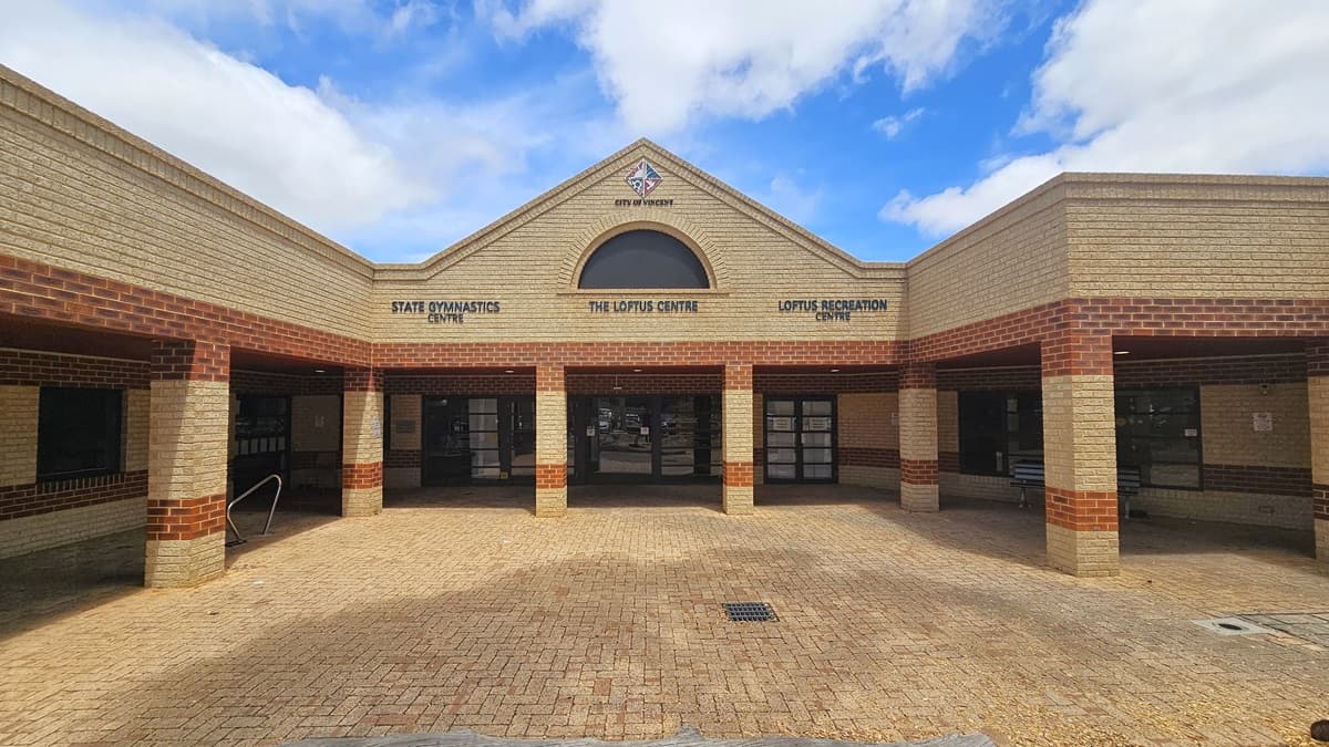 A low-set structure constructed with brown and cream bricks, with the building concourse sharing the brown brick pattern. The facility banner hangs on the left side. The parking area directly adjoins the building, hosting cars of various models and colors. Humps on the road are marked by yellow and black lines. A step-free entrance with a ramp is positioned in the middle. Lamp posts and trees are also visible in front of the building.