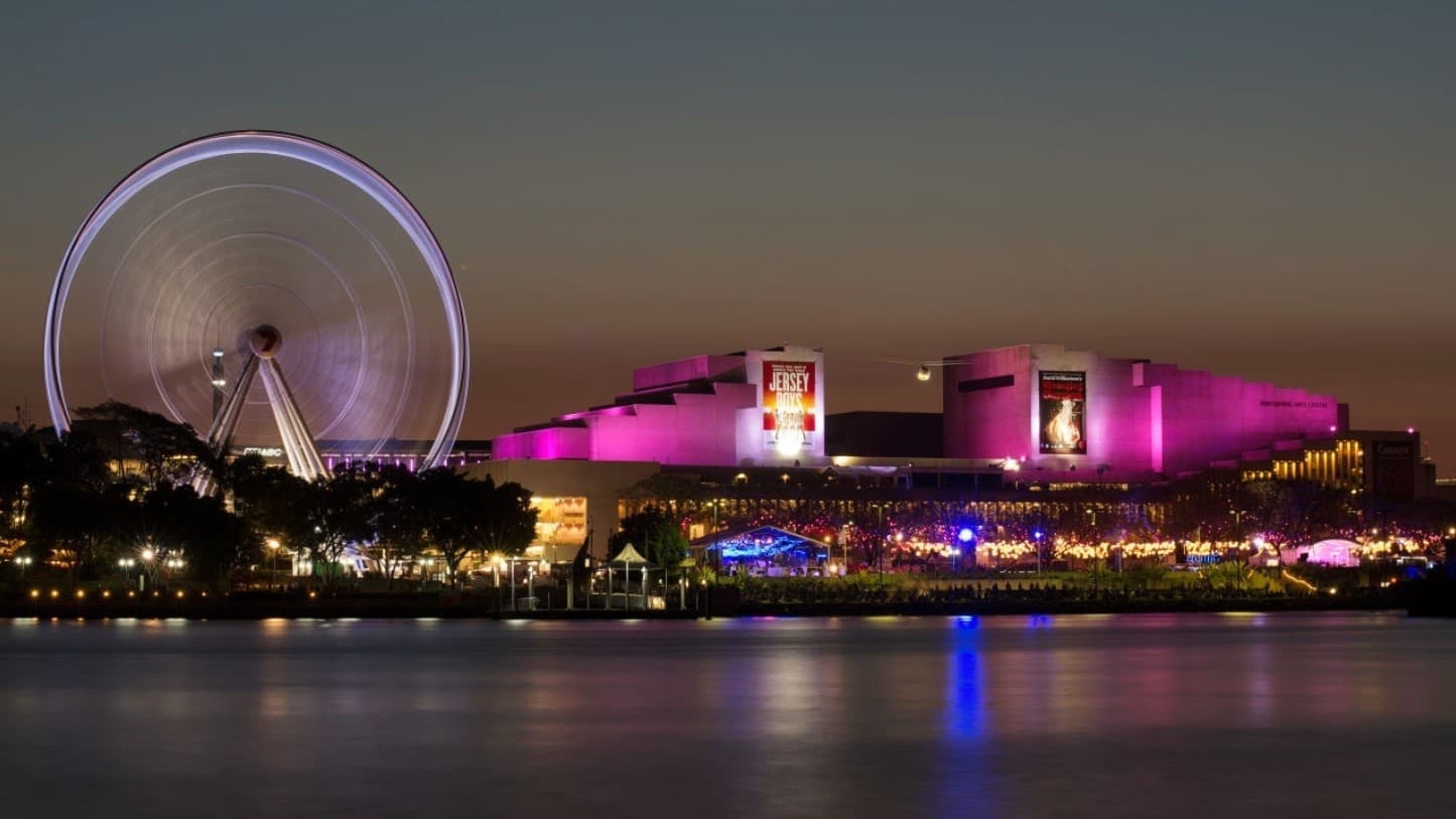 A night-time view of the Queensland Performing Arts Centre with illuminated red lighting. Large advert banners are affixed to the building's front. To the left, The Wheel of Brisbane is captured in motion with blurred lights, suggesting movement. The street is crowded with lights. The sky is dusk-toned, and reflective water is in the foreground.