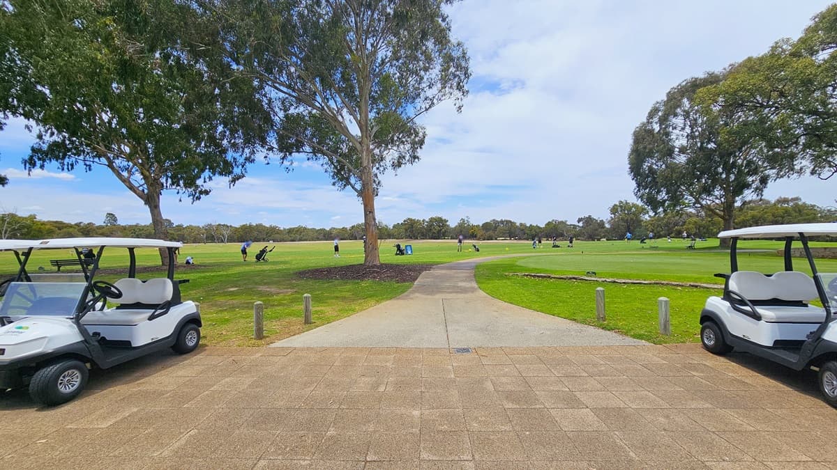Golf carts are arranged in a row near the sloping grassy area on the left, bordered by concrete bollards. In the middle, there is a low-rise building with a veranda where golf bags are visible. Picnic tables and chairs are arranged under umbrella shades in front of the building. The front of the building is covered with green grass, and tall trees surround the area.