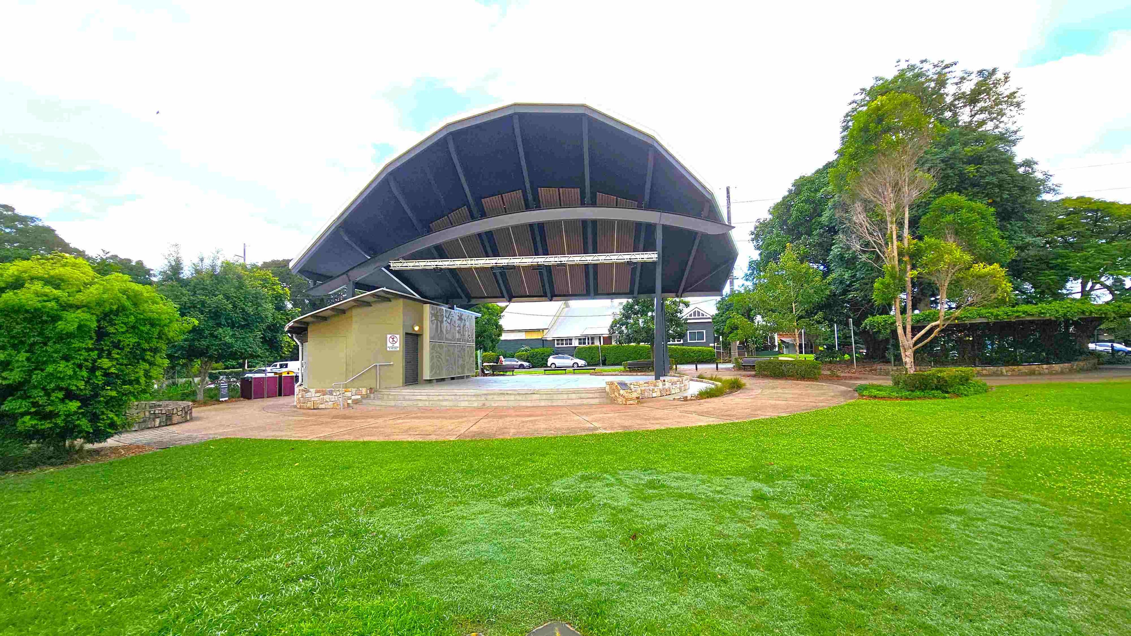 A large, open-air stage with a curved roof is in the centre, surrounded by a grassy area. The stage has a beige building attached to it on the left, with steps leading up. Trees and shrubs are on both sides, with a few small trees on the right. In the background, there are residential houses and a parked car. The ground is a mix of grass and paved pathways.