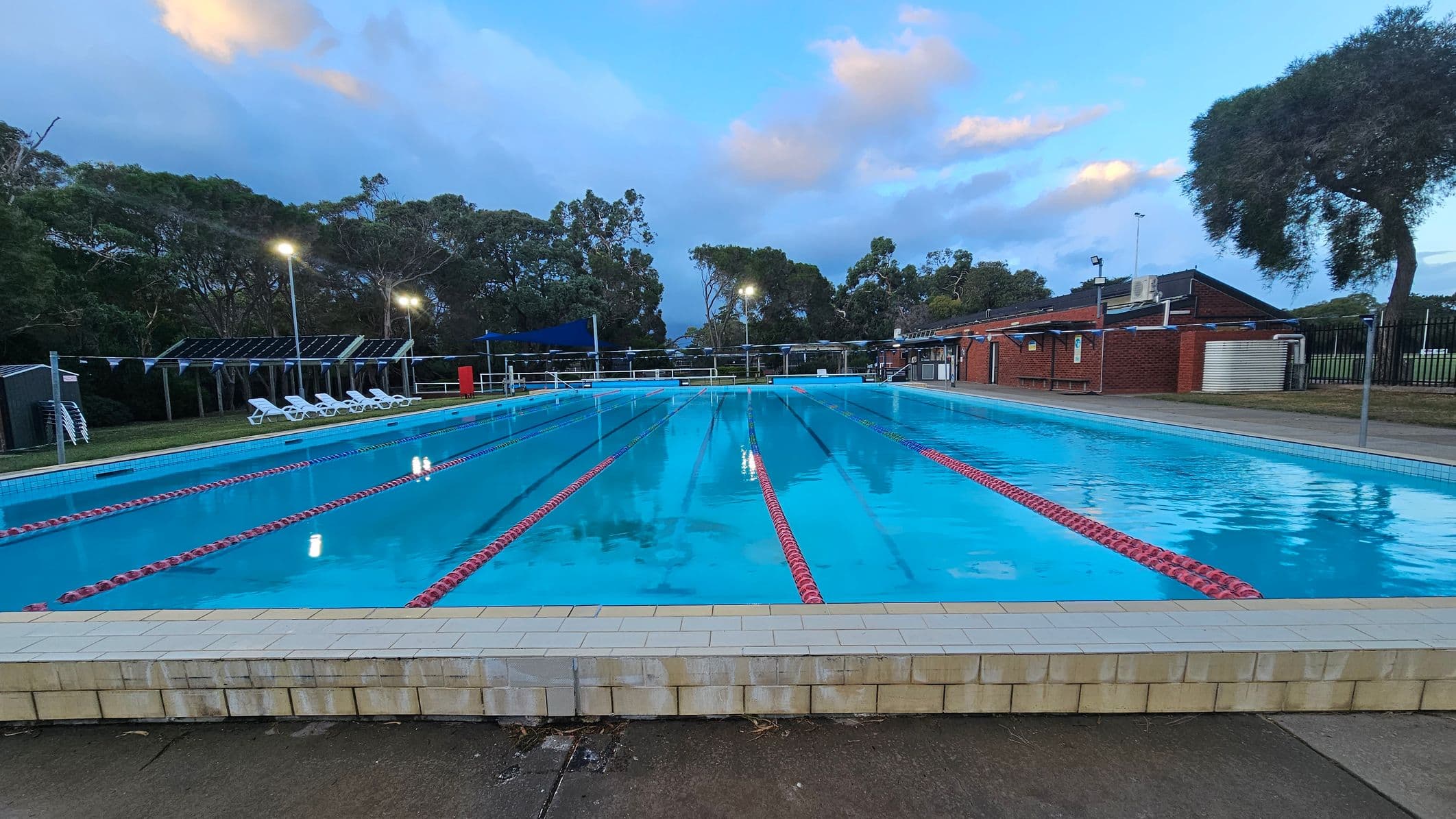 An outdoor swimming pool with clear blue water is in the centre, surrounded by a tiled edge. On the left, there is a grassy area with white lounge chairs and a blue shade sail. To the right, a concrete pathway runs alongside the pool. In the background, a brick building is visible, along with tall trees and a fence. The pool has lane dividers, and there are metal handrails at the pool's edge. The area is well-lit with tall lights.