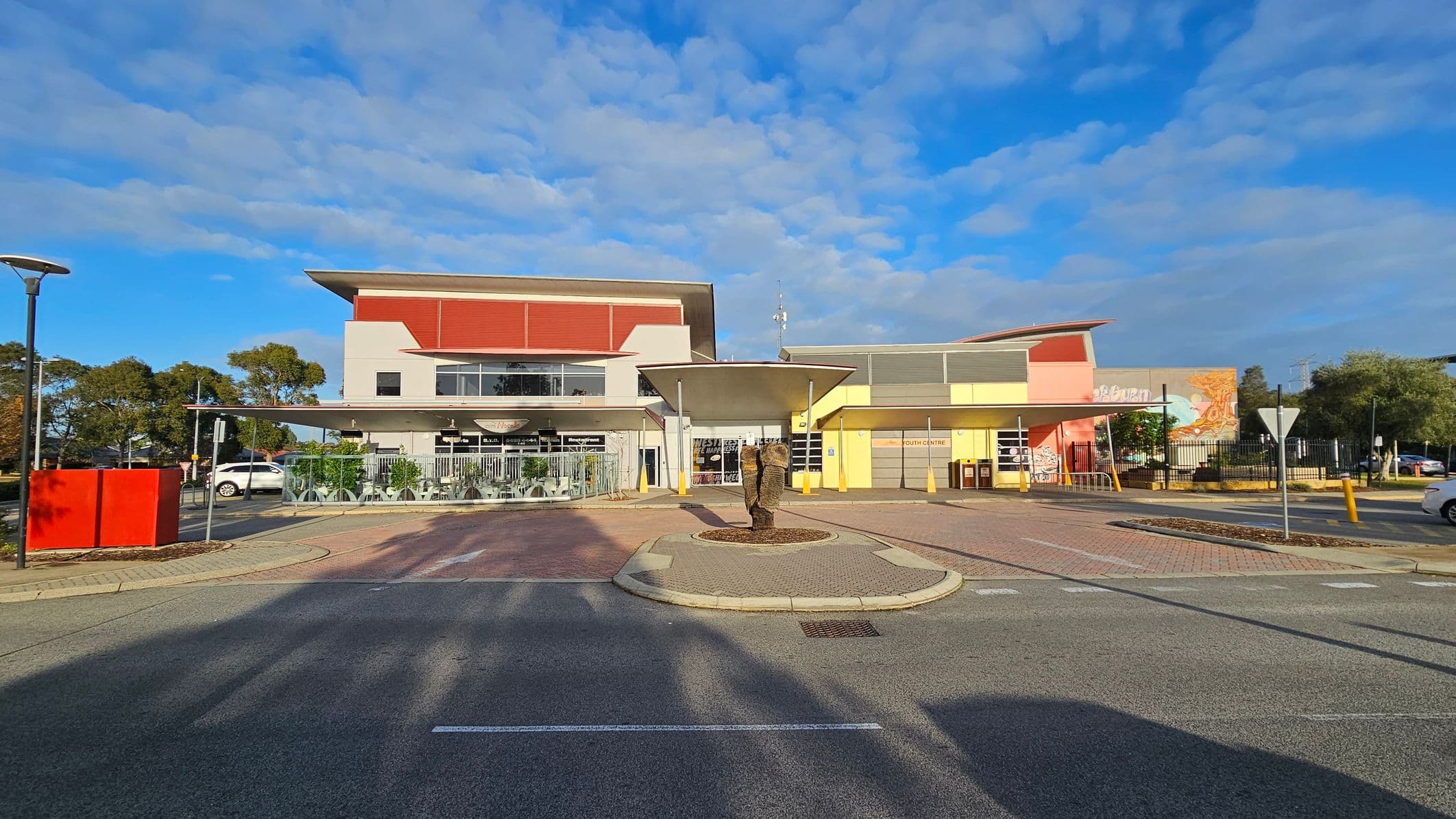 A modern building with a flat roof and a mix of red, yellow, and grey colours. In the centre foreground, there's a small roundabout with a sculpture. The left side has a glass-enclosed outdoor seating area with tables and chairs. On the right, a yellow section of the building with a sign reading "Youth Centre" and a mural. The ground is paved with a mix of asphalt and brick patterns. Trees and parked cars are visible in the background.