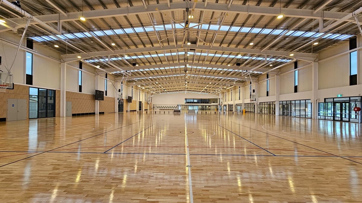 To the right is the stadium's spectator area with two rows of folded chairs for seating. A glass railing secures the area. To the left is the stadium, featuring timber floors marked for various ball games and an industrial ceiling with exposed beams, skylights, and aluminum insulation.