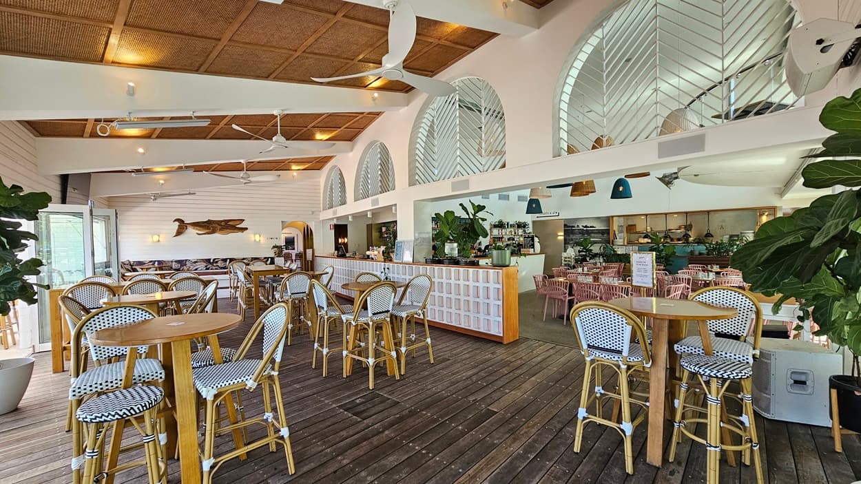 On the left, is the entrance to the main bar in the centre are high tables and bar stools and to the right are low set tables and chairs under striped umbrellas. The road is further to the right and gardens separate the road from the outdoor dining area. The sun is shining through the umbrellas and trees.