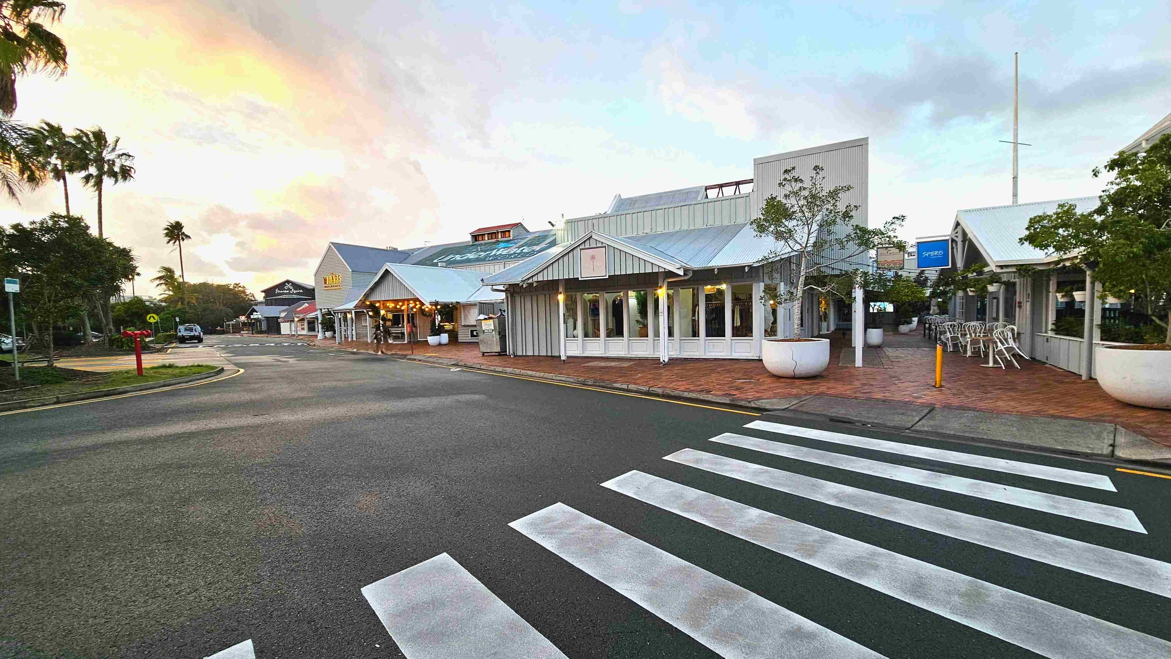 A street scene featuring a pedestrian zebra crossing leading to a complex of low-rise buildings with corrugated metal roofs and large windows. The buildings are surrounded by brick paving and large white planters with small trees. Outdoor seating with stacked chairs is visible. A sign reads 'Wharf' on one building. The area is open with palm trees in the background and a few parked vehicles.