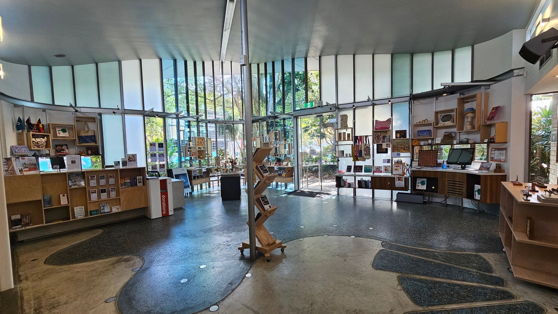 A spacious room with a curved wall of tall windows in the background, allowing natural light to illuminate the space. The floor is a mix of polished concrete and textured surfaces. On the left, there are wooden shelves displaying books and brochures. In the centre, a zigzag-shaped wooden book stand is positioned. To the right, more shelves hold various items, including bags and artwork. The ceiling is smooth and light-coloured, complementing the modern aesthetic.
