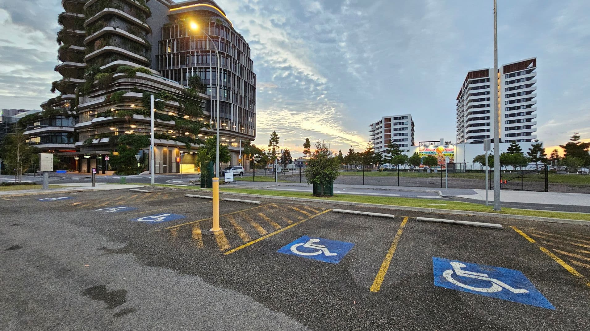 In the foreground, there are parking spaces marked with blue disabled signs. A yellow bollard stands near the centre. On the left, a modern building with curved balconies and greenery is visible. In the background, two white multi-storey buildings are on the right. Trees and a billboard are situated between the buildings. The ground is asphalt, and a streetlamp is lit, casting a warm glow.