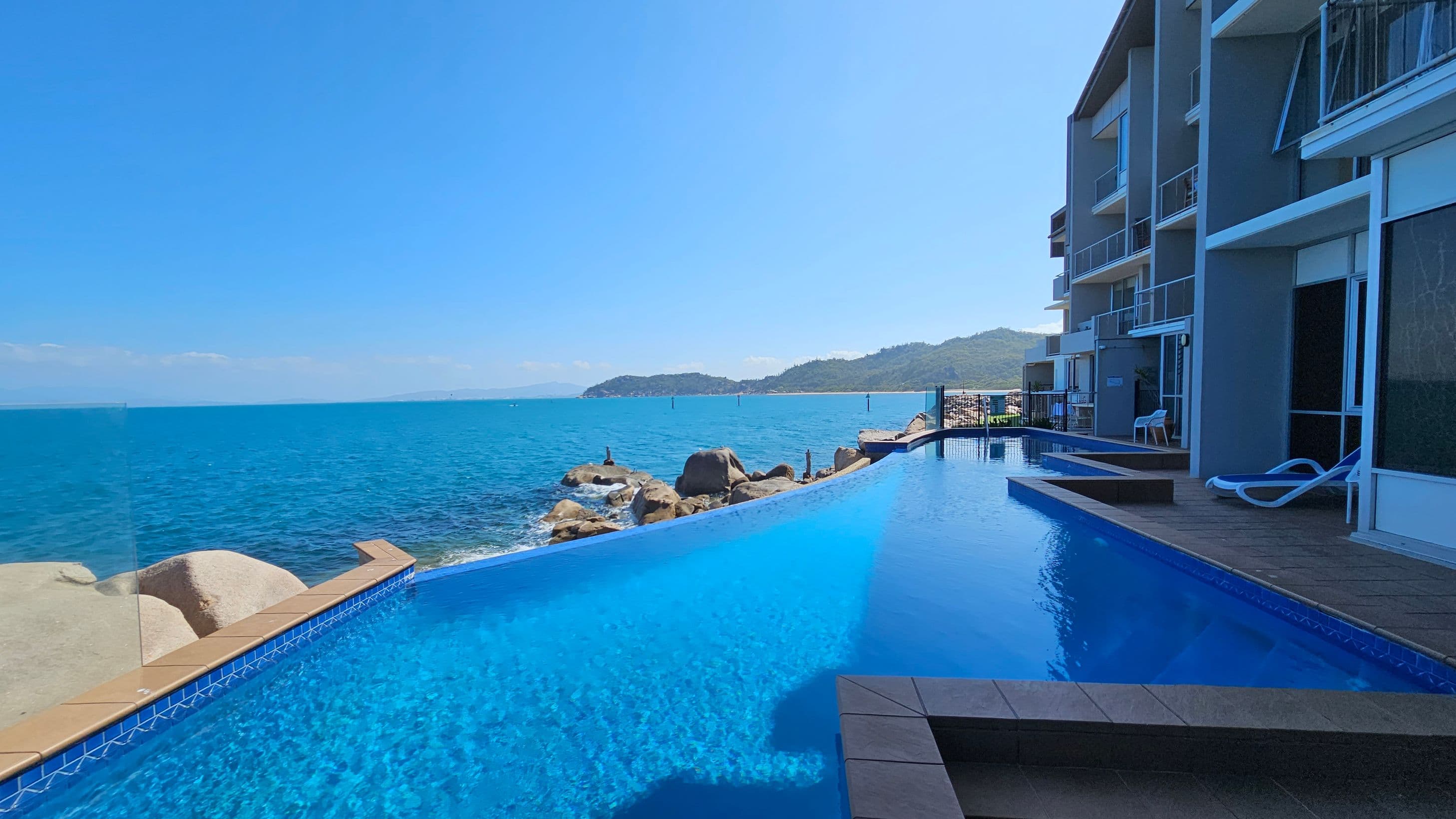 An infinity pool is in the foreground, with clear blue water extending towards the ocean. On the right, a modern building with balconies overlooks the pool, and a couple of white sun loungers are placed on the deck. The background features a rocky shoreline and a distant view of a hilly landscape across the water. The sky is clear and blue.