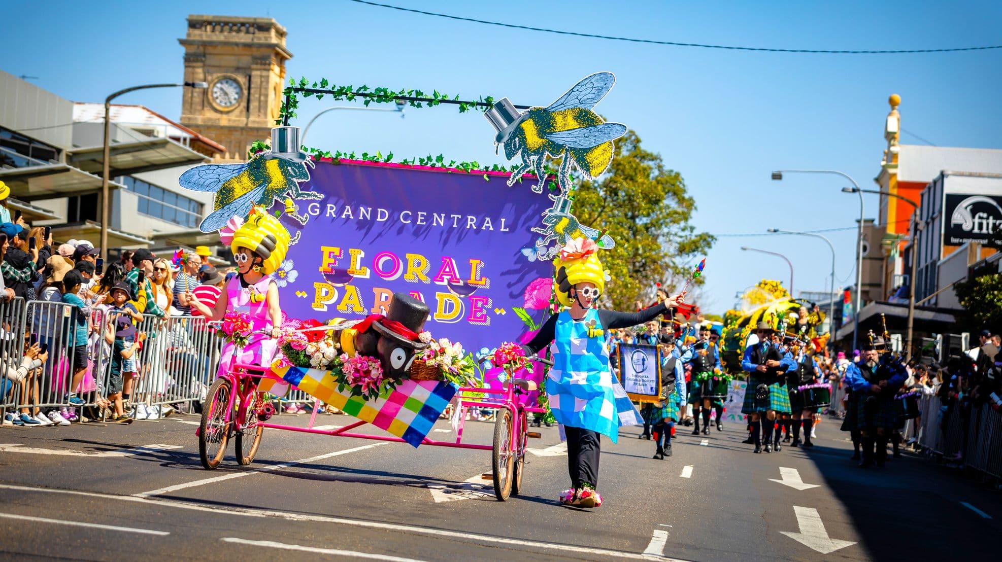 A vibrant parade scene with participants in colourful costumes pushing a decorated cart. The cart features a large bee and floral arrangements, with a banner reading "Grand Central Floral Parade". Spectators line both sides of the street behind metal barriers, capturing the event on their phones. In the background, a clock tower and various buildings are visible. The street is filled with other parade participants, including a group of bagpipers.