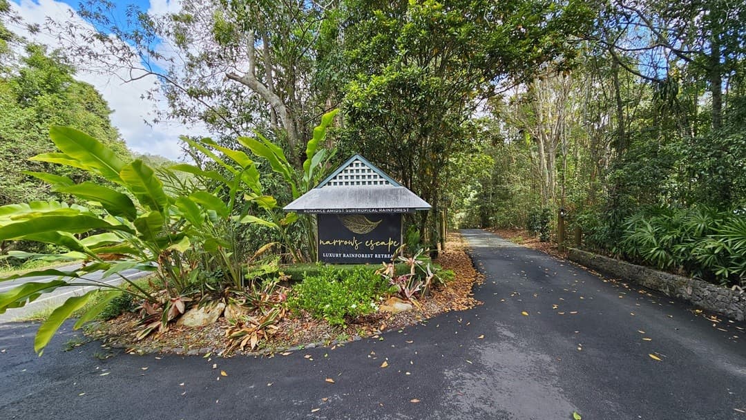 Arriving at Narrow Escape, you'll encounter a distinctive sign proudly displaying "Narrows Escape Luxury Rainforest Retreat" at the edge of a meandering road leading to the retreat. The sign, designed to resemble a small house, is situated within a landscaped area adorned with tall banana fig plants and other ornamental greenery. The pathway is gracefully bordered by shrubs and trees, welcoming you to the retreat.