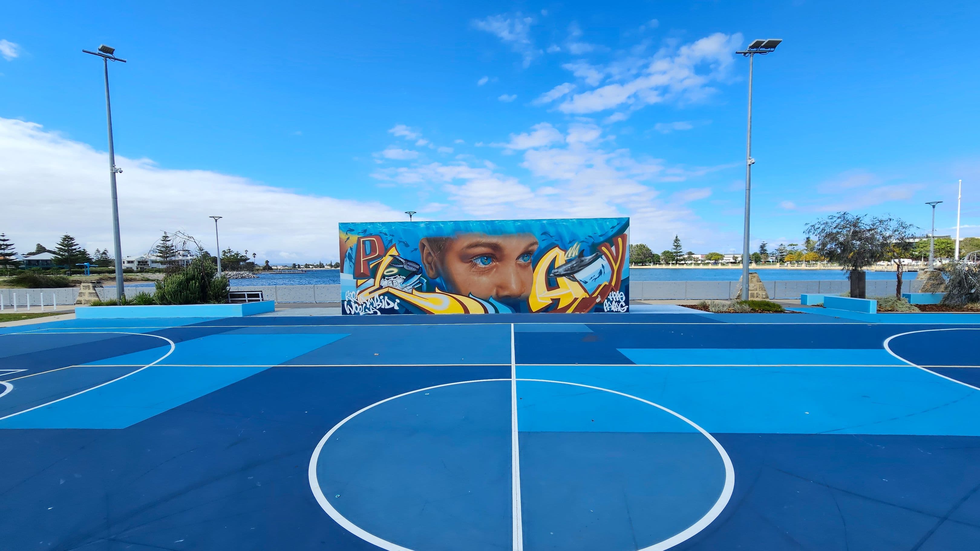 A vibrant outdoor basketball court with various shades of blue on the floor. In the centre, a large mural features a face with blue eyes, surrounded by colourful graffiti. The background shows a body of water with trees and buildings along the shore. Tall streetlights are positioned on either side of the court, and there are benches and greenery around the perimeter.