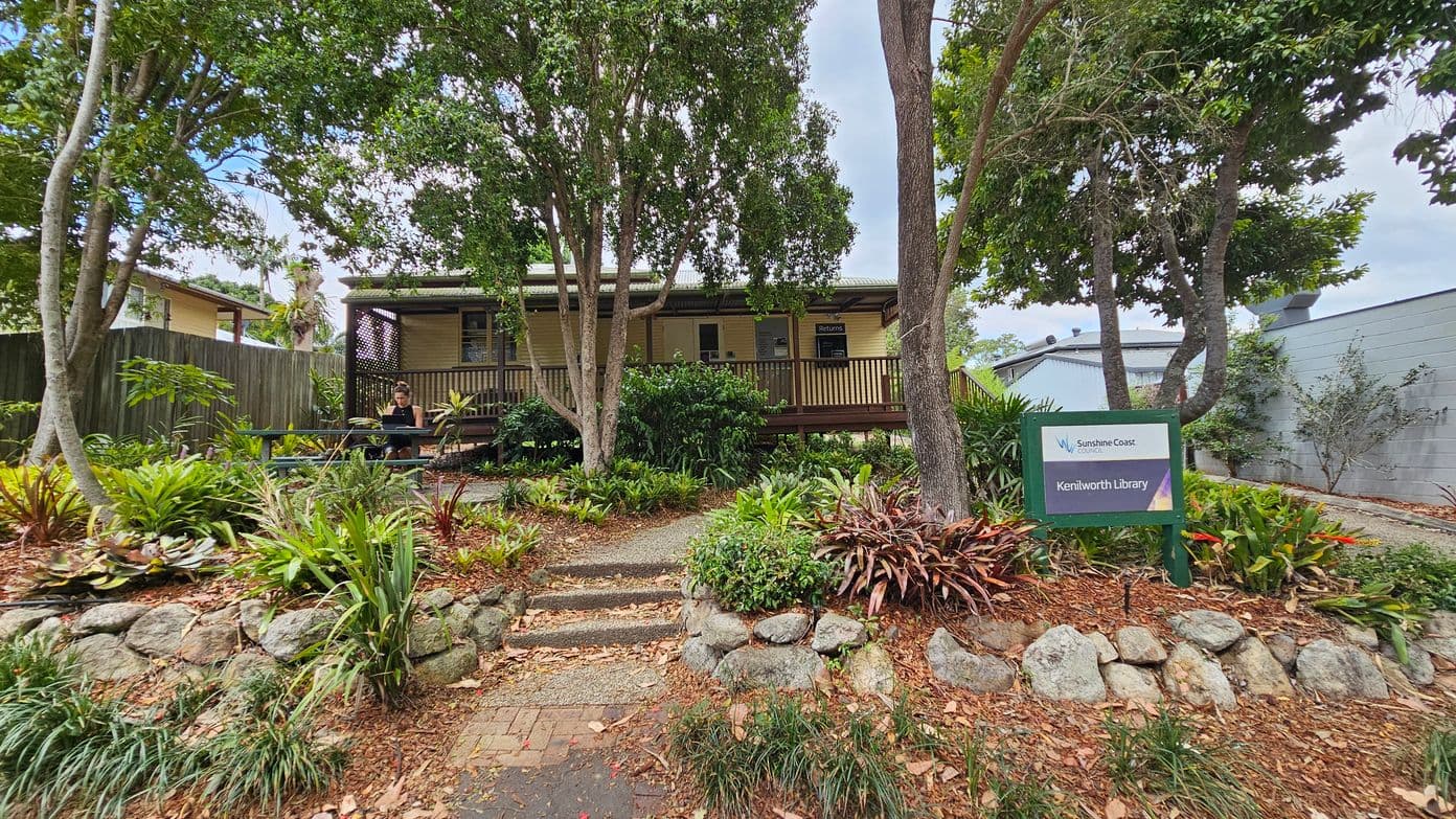 A small library building is partially visible in the background, surrounded by lush greenery and trees. In the foreground, a sign reads "Kenilworth Library." A person sits at a picnic table to the left, under the shade of the trees. A gravel driveway leads to the right, where a car is parked near a grey building. The area is landscaped with rocks and various plants, creating a serene, natural setting.