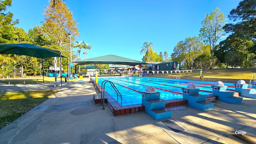 View from the diving platform of the 25m, 6-lane outdoor pool at Dayboro Pool and Gym. The pool deck has a rough surface floor to help avoid slipping.  Each lane has a two-step stair with a grey non-slip cover blue diving platform. On top of the diving platform is a yellow semi-circle with a red caution on it. There is a green sunshade on the left with grey metal benches. Yellow floating devices are wrapped around poles with strings of blue triangular flags.