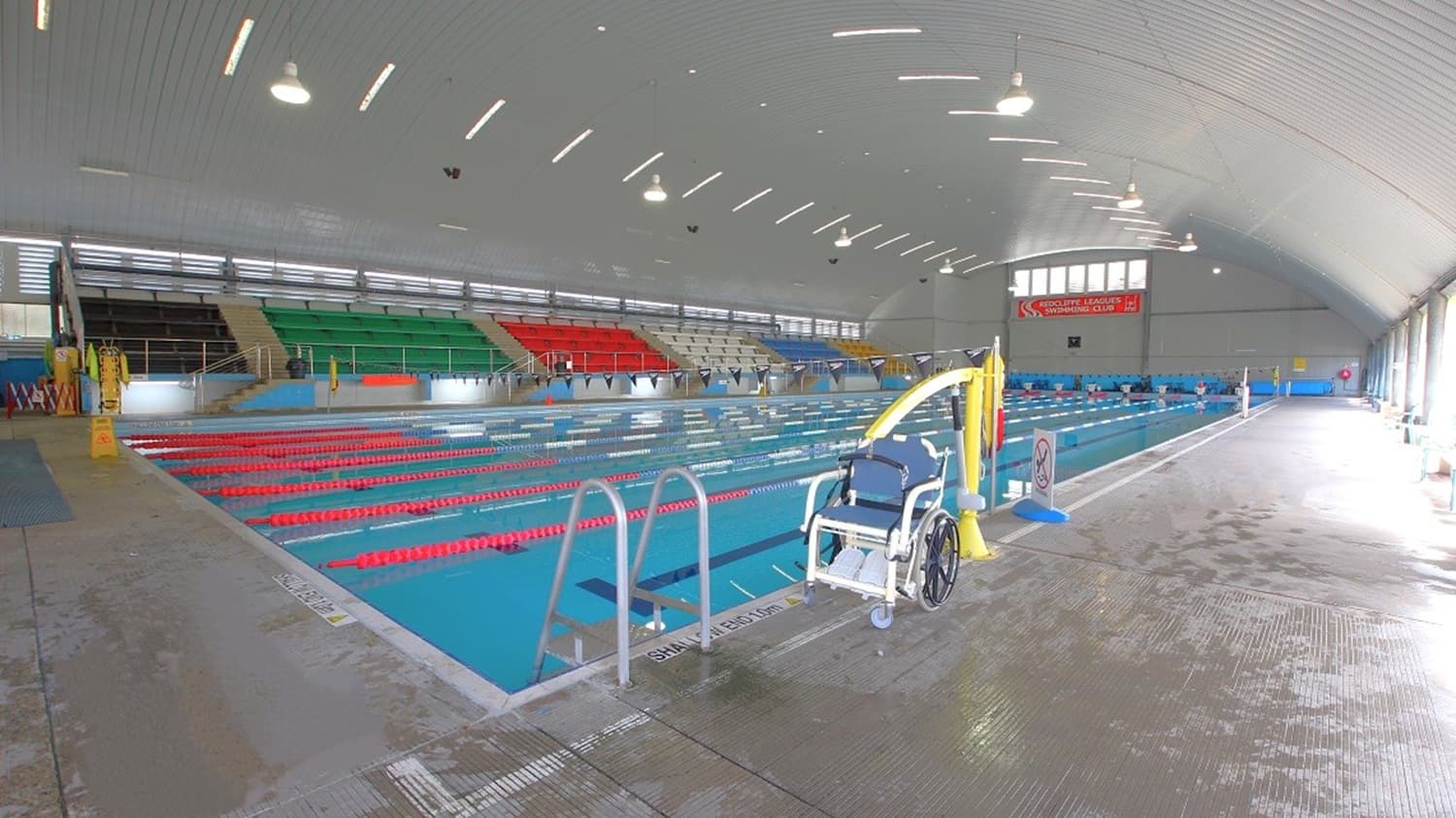 An indoor swimming pool with multiple lanes marked by red lane dividers. On the left, there are tiered seating areas with green, red, and blue seats. On the right, a wheelchair lift is positioned by the poolside. The ceiling is curved with evenly spaced lights, and the floor is wet and textured for safety. Large windows on the right allow natural light to enter.