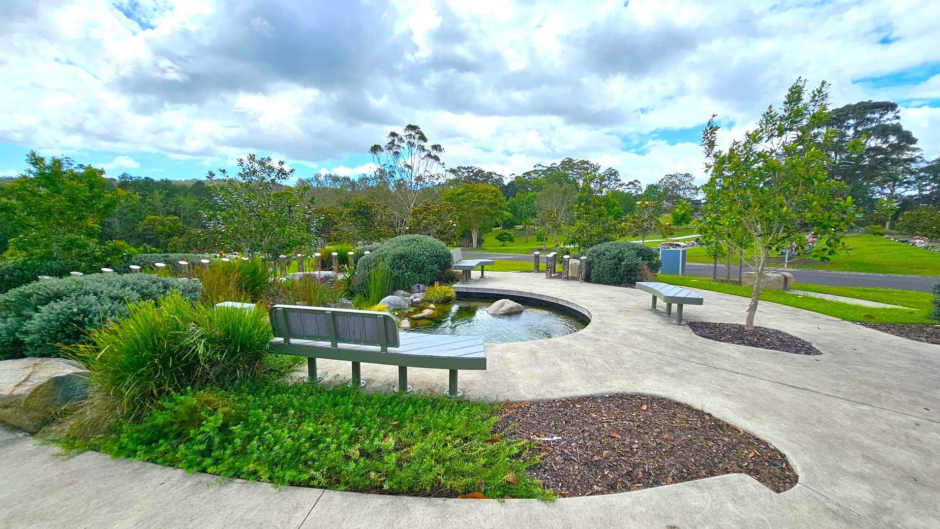 A landscaped garden with a curved concrete path and a small pond in the centre. On the left, there are tall palm trees lined up, and a few benches are placed along the path. Bushes and various plants are scattered throughout the area. On the right, decorative metal posts line the path, and more greenery extends into the distance. The background features a wooded area with dense trees.