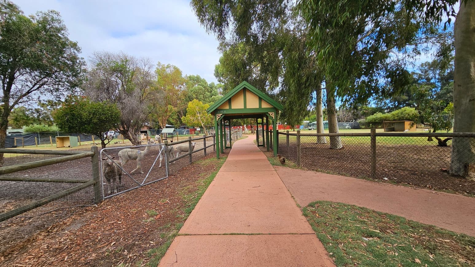 A pathway with a reddish surface leads through a small green-roofed shelter in the centre. On the left, there are sheep behind a wire fence, with trees and a small wooden structure in the background. On the right, more trees and a fenced area with a small shed are visible. The ground is a mix of grass and dirt, and the area is surrounded by various trees.
