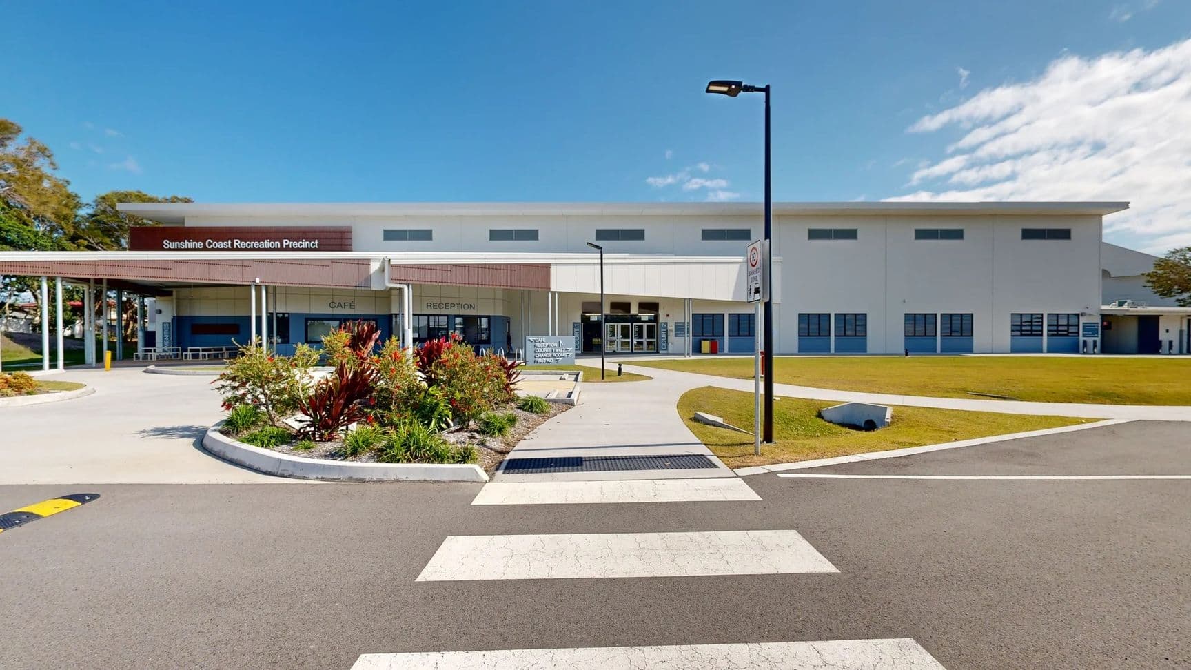A large building with a flat roof is at the centre, featuring large glass doors and signage indicating "Reception" and "Café." On the left, there is a shaded outdoor seating area supported by tall, grey columns. Adjacent to this, a designated parking spot for disabled access is marked with a blue symbol, alongside yellow striped lines indicating reserved spaces. To the right, a sign points towards various facilities like "Court 1" and "First Aid." The floor is smooth concrete.