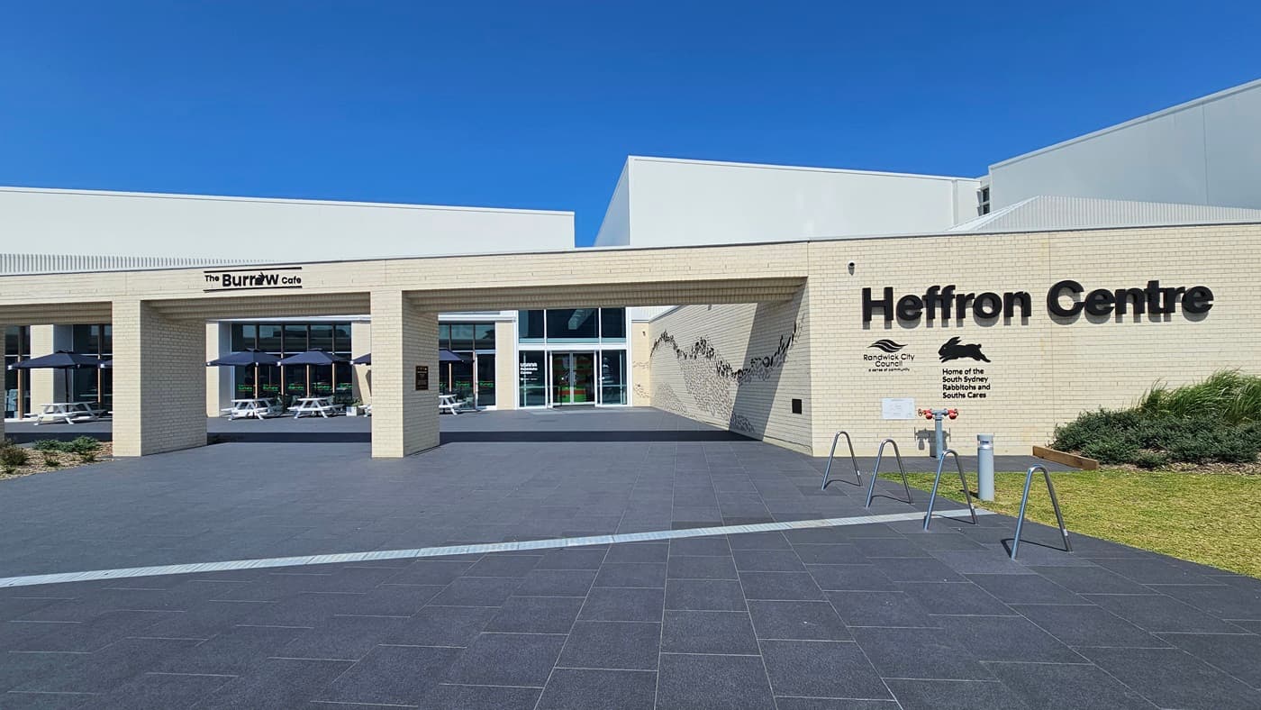 The building features a spacious front with a brick wall facade, displaying the facility's name and its partners. On the right, there's a landscape with grass and ornamental plants. Next to the wall is the accessible parking area, equipped with signs and concrete bollards. Behind it stands a two-story modern structure with glass walls and corrugated roofing. Tables and chairs are arranged under large umbrella shades. A pedestrian crossing extends from the car park.