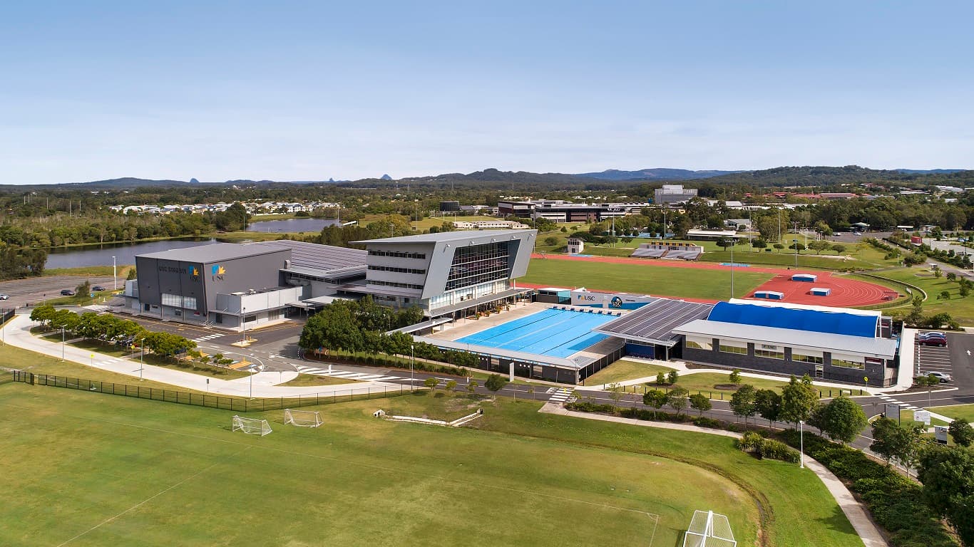 The USC Aquatic & Fitness facility is located between two sporting fields. It’s a prominent building with a tree-lined frontage and 3 unique architectural styles. The facility includes an open pool and a covered pool that has a distinctive tunnel-like shade, lending it a serene and charming appearance. The left wing is a low-rise square structure with grey patterns. The right-wing is a four-story grey building with a slanted design made of metal and glass.