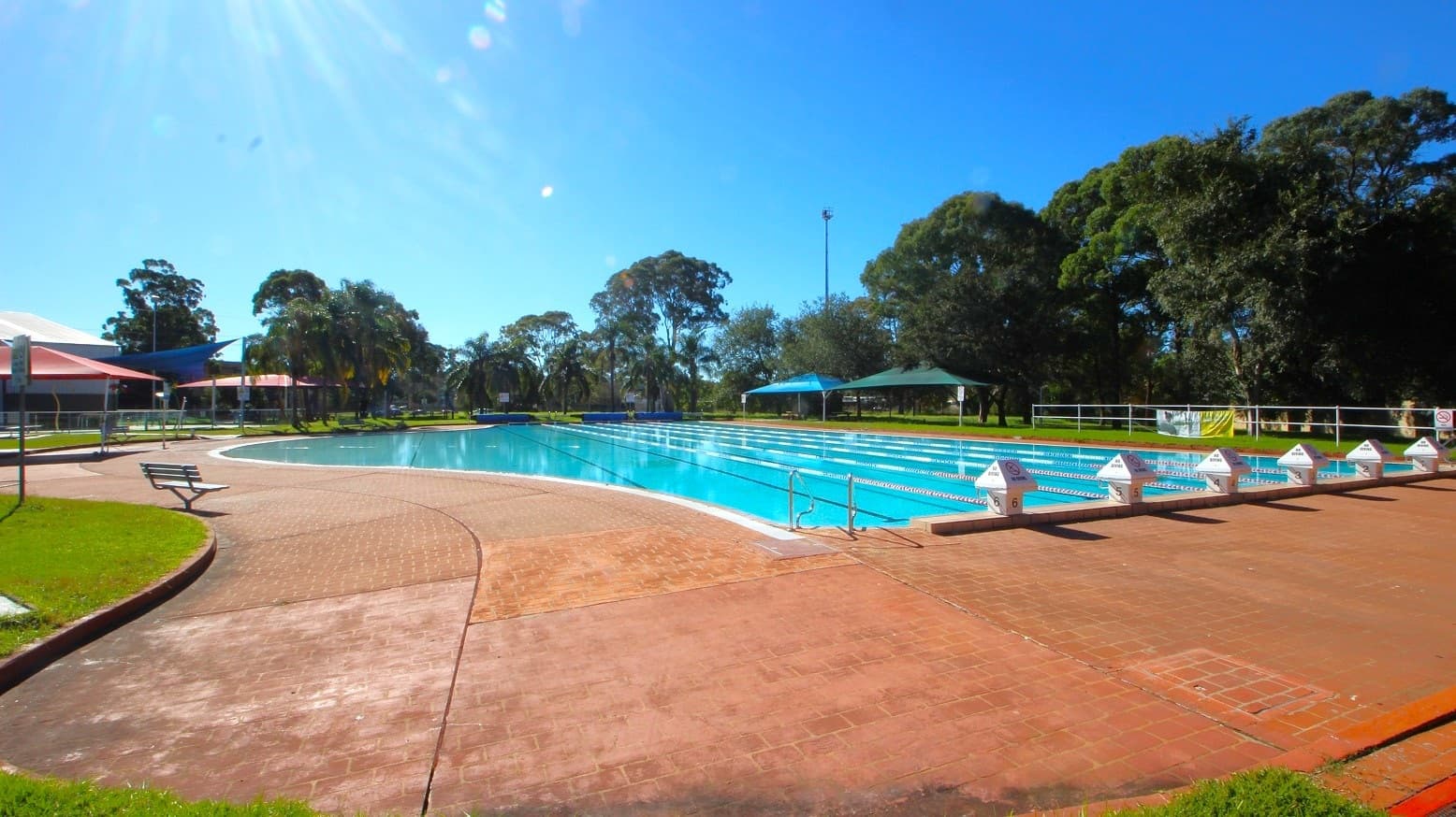 External pool with a curvy side at Michael Wendon Aquatic Leisure Centre. The pool has 6 lanes separated by white and blue floating ropes. Intersecting horizontally to the pool is a string of blue flags hanging in the air above the pool. Surrounding the pool is a brick pathway.