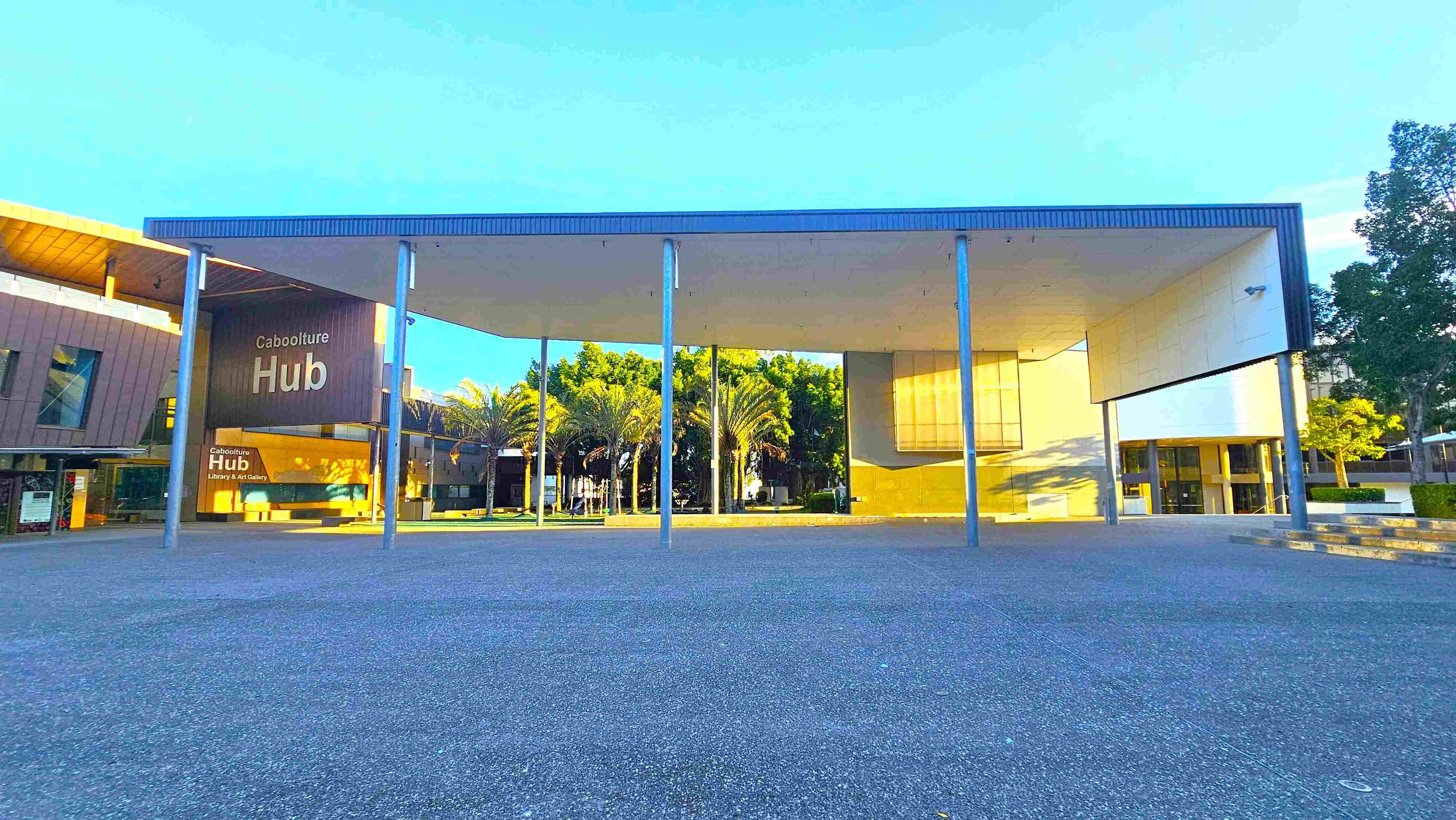 A modern building with a large, flat roof supported by metal columns. On the left, a sign reads "Caboolture Hub" with "Library & Art Gallery" below it. The building has a mix of glass and wooden panels. In the centre, palm trees are visible behind the structure. On the right, there is a continuation of the building with more glass and a small set of steps. The ground is paved with grey concrete, and there are trees in the background.