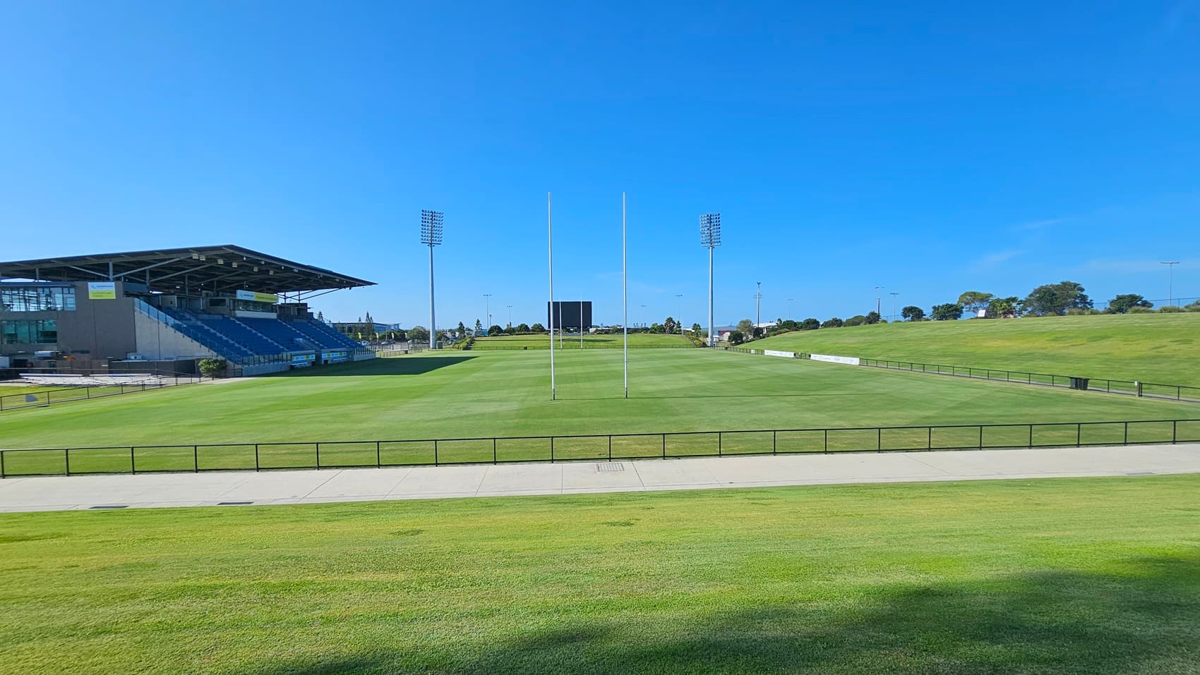 A rugby field with green grass and goalposts in the centre. On the left, there is a covered grandstand with blue seating. In the background, tall floodlights and a scoreboard are visible. The field is surrounded by a low fence, and there is a paved walkway in the foreground. Trees and open grassy areas are in the distance.