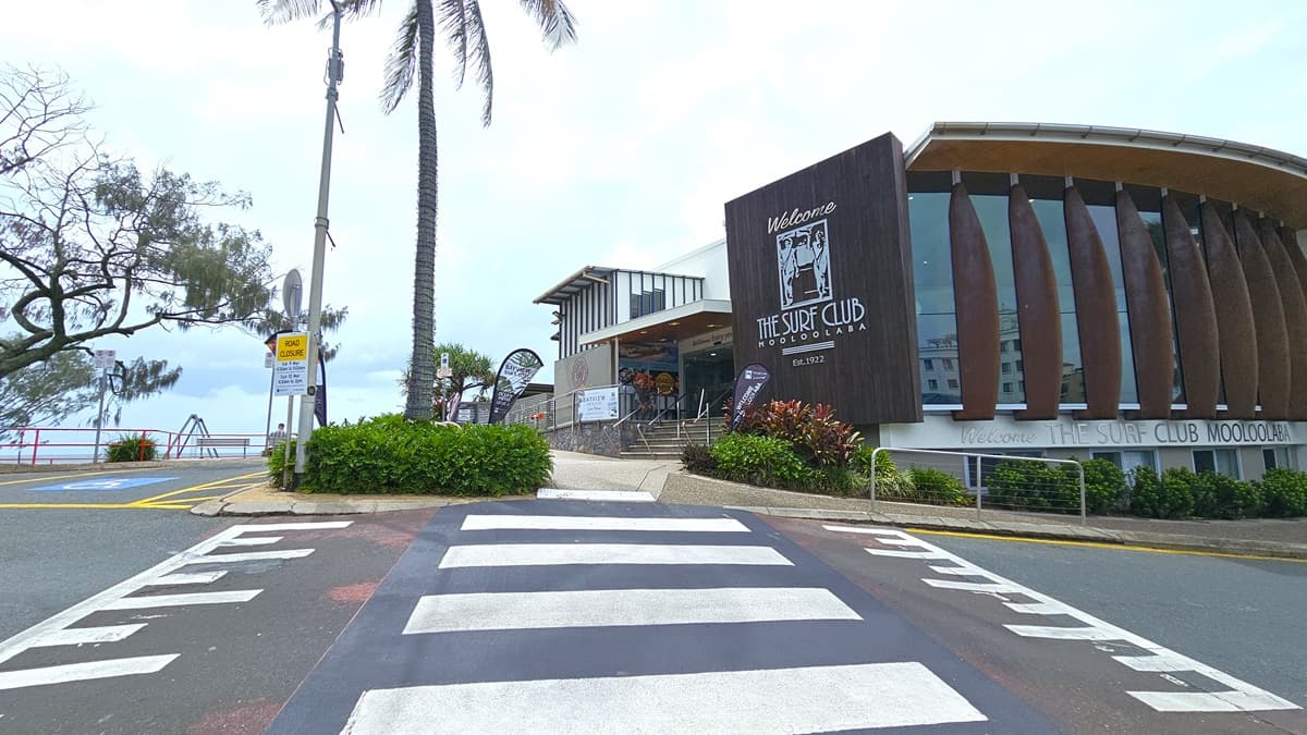 A wide-angle view of the entrance to a modern building labelled "Mooloolaba." A flight of stairs with handrails and truncated domes leads up to glass double doors, flanked by stone walls. A ramp with railings is on the left. A wall-mounted sign reads 'Welcome, enjoy your visit!' Below it, a banner displays 'BAYVIEW Bar & Bites'. Palm trees are visible on the left.
