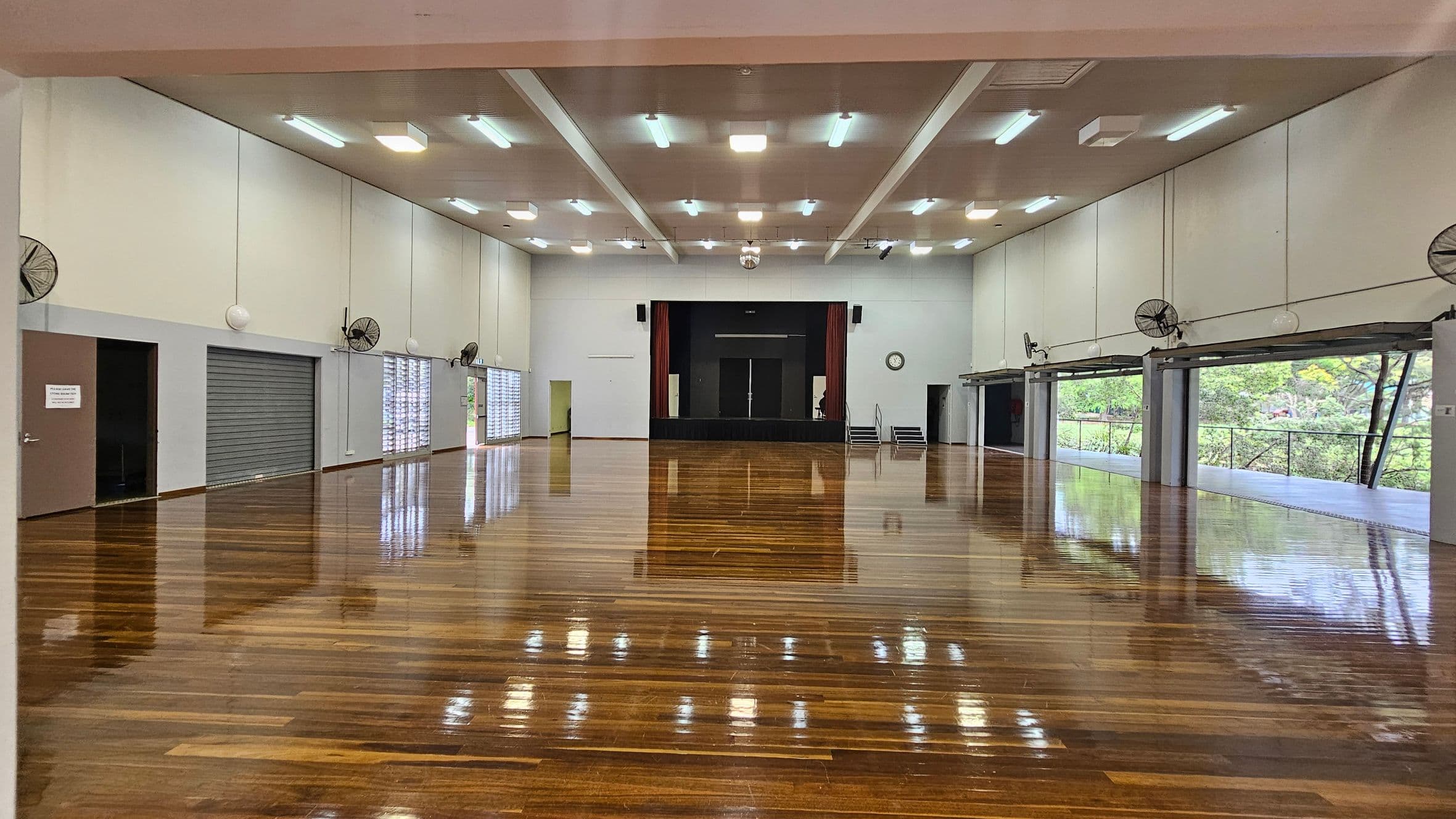 A spacious hall with a polished wooden floor and a high ceiling featuring fluorescent lights. On the left, there are closed roller shutters and a door. On the right, large windows with open shutters reveal greenery outside. Ceiling fans are mounted along the walls. At the far end, a stage with red curtains and a black backdrop is centred, with a clock on the wall to the right.