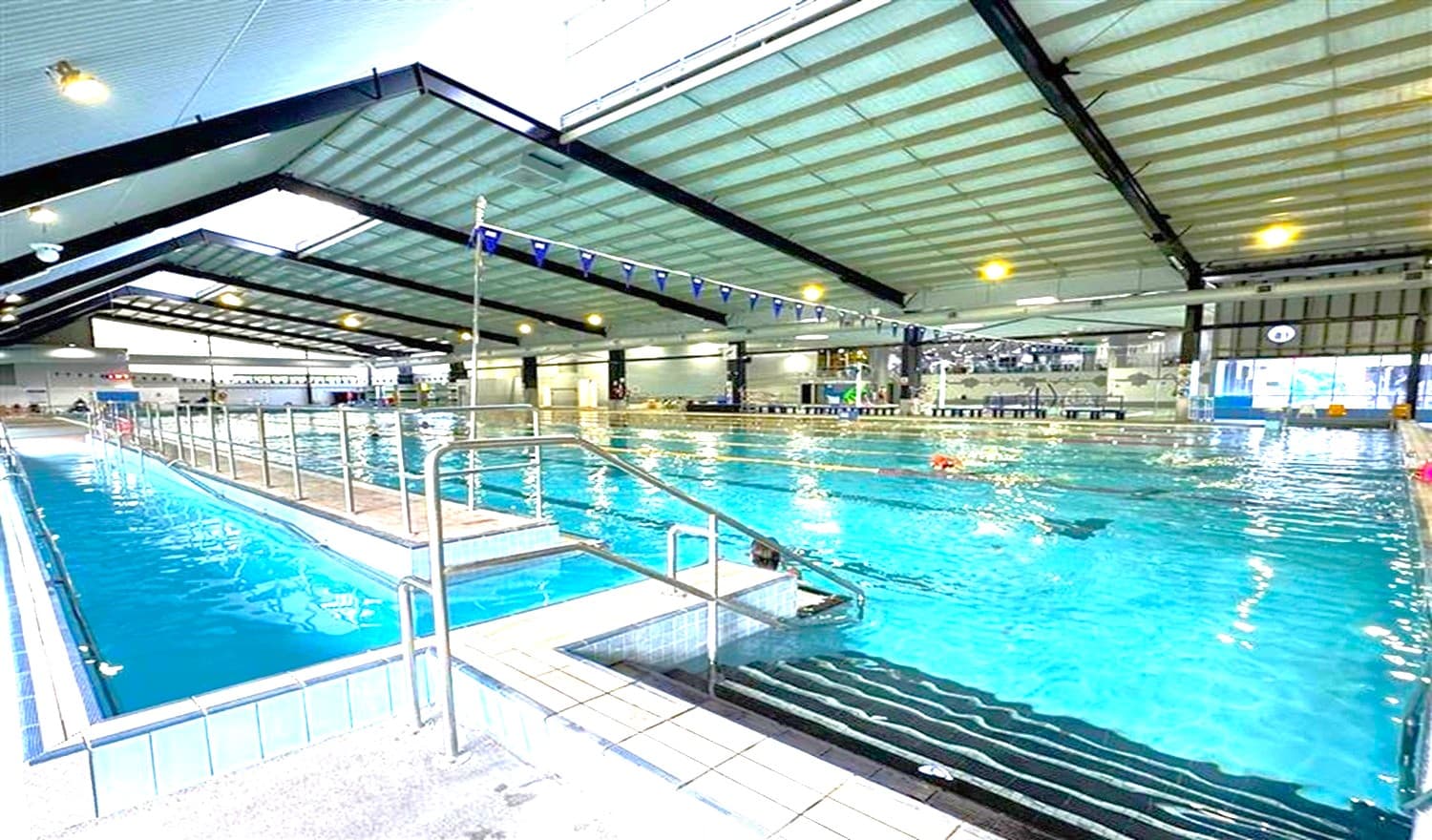 Indoor swimming pool with a high, sloped ceiling featuring exposed beams. On the left, a narrow lane with metal railings and steps leads into the water. The main pool area is on the right, with several swimmers in lanes. The floor is tiled, and the ceiling has large lights. In the background, there are windows and a seating area.