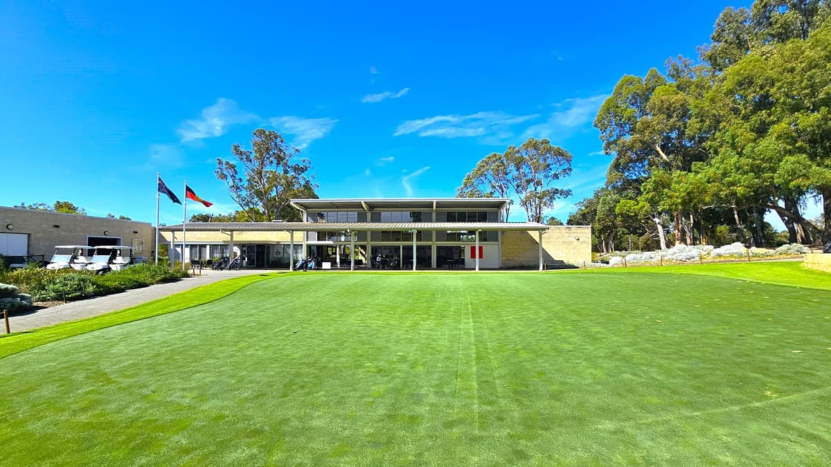 An expansive green space featuring well-maintained grass and synthetic turf. To the left, an ascending path leads to the building. In the middle stands a two-story cream building. Two flags are positioned on the right, accompanied by three golf carts. Shrubs and tall native trees encircle the area.