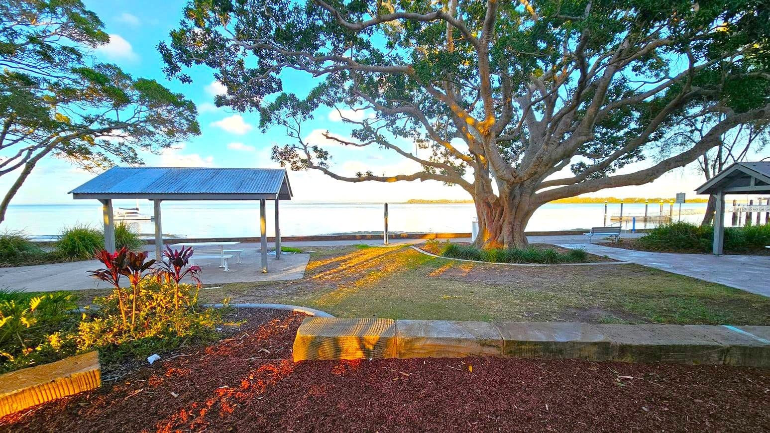 A large tree with sprawling branches dominates the centre, casting shadows on the ground. To the left, a small shelter with a corrugated metal roof stands near the water's edge. On the right, another similar shelter is partially visible. The foreground features a landscaped area with red mulch and plants. The background shows a calm body of water with a boat and a distant shoreline. The ground is a mix of grass and paved paths.