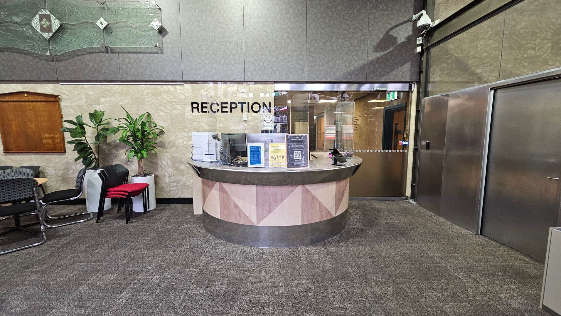 A reception area with a curved desk in the centre, featuring informational displays and a transparent screen. The word "RECEPTION" is visible on the wall behind. To the left, there are several chairs, a wooden plaque, and two potted plants. On the right, there are metallic doors and a security camera above. The floor is carpeted in a grey pattern, and the walls have a textured design with a decorative glass panel above.