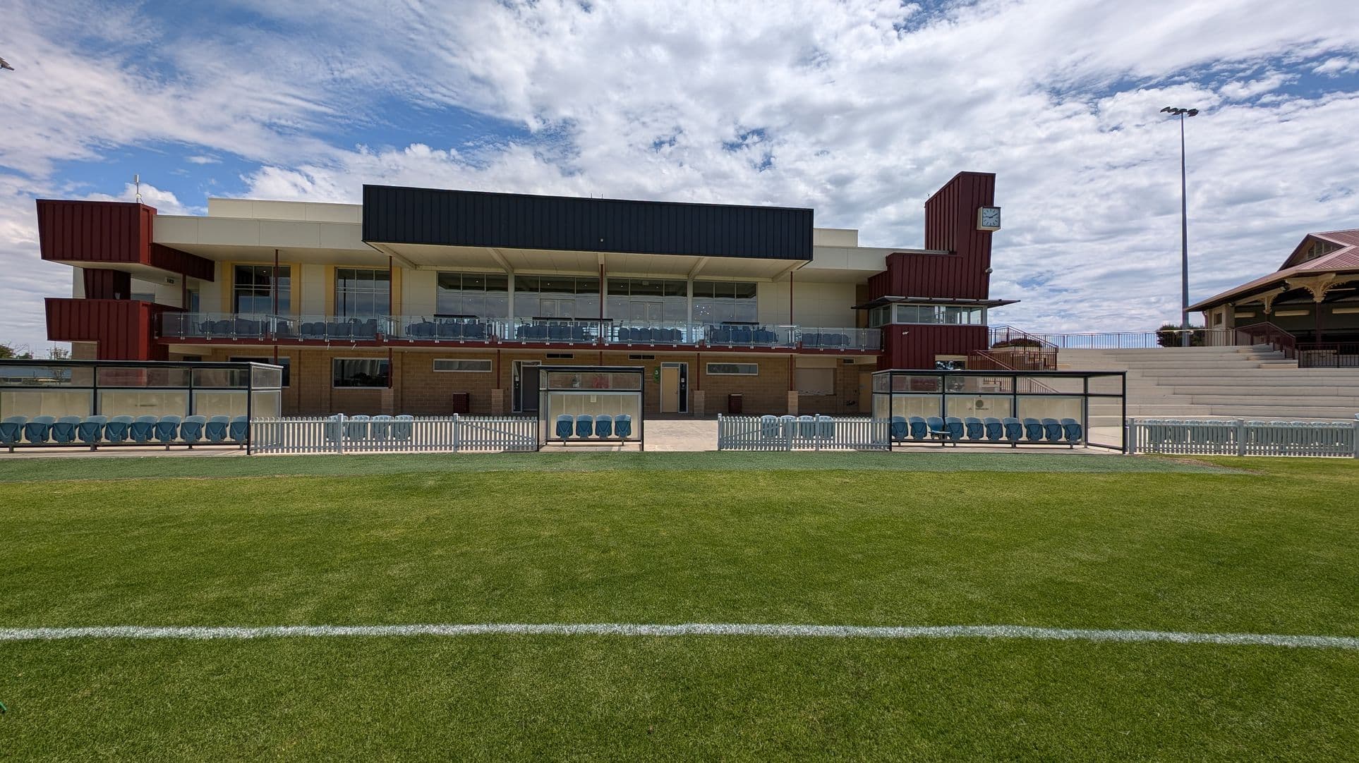 A modern building with a glass facade on the left, featuring horizontal slats and a protruding entrance. The sign reads "Port Pirie Sports Precinct" and "Memorial Oval". The right side has a beige brick wall with a small circular window. In the background, there is a sports field with trees and a tall chimney. The foreground shows a concrete pathway with some shrubs on the right.