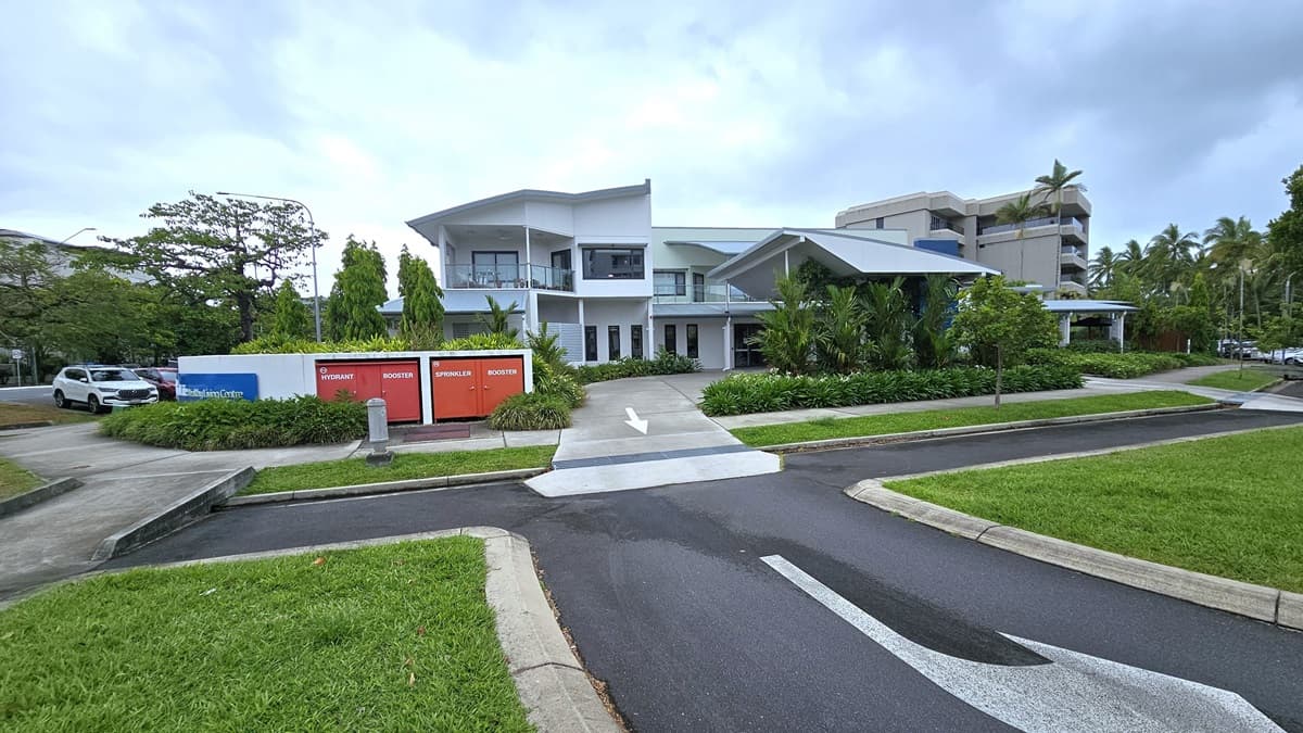 On the left are parked vehicles on the side of the road. A concrete pathway extends from the car park to the entrance. In the centre stands a white concrete fence displaying the facility logo "Spinal Life Healthy Living Cairns". Behind it, a modern two-story building crafted from glass and concrete is nestled amidst trees, landscaped ornamental plants, and green grass. Step-free ramps are accessible.