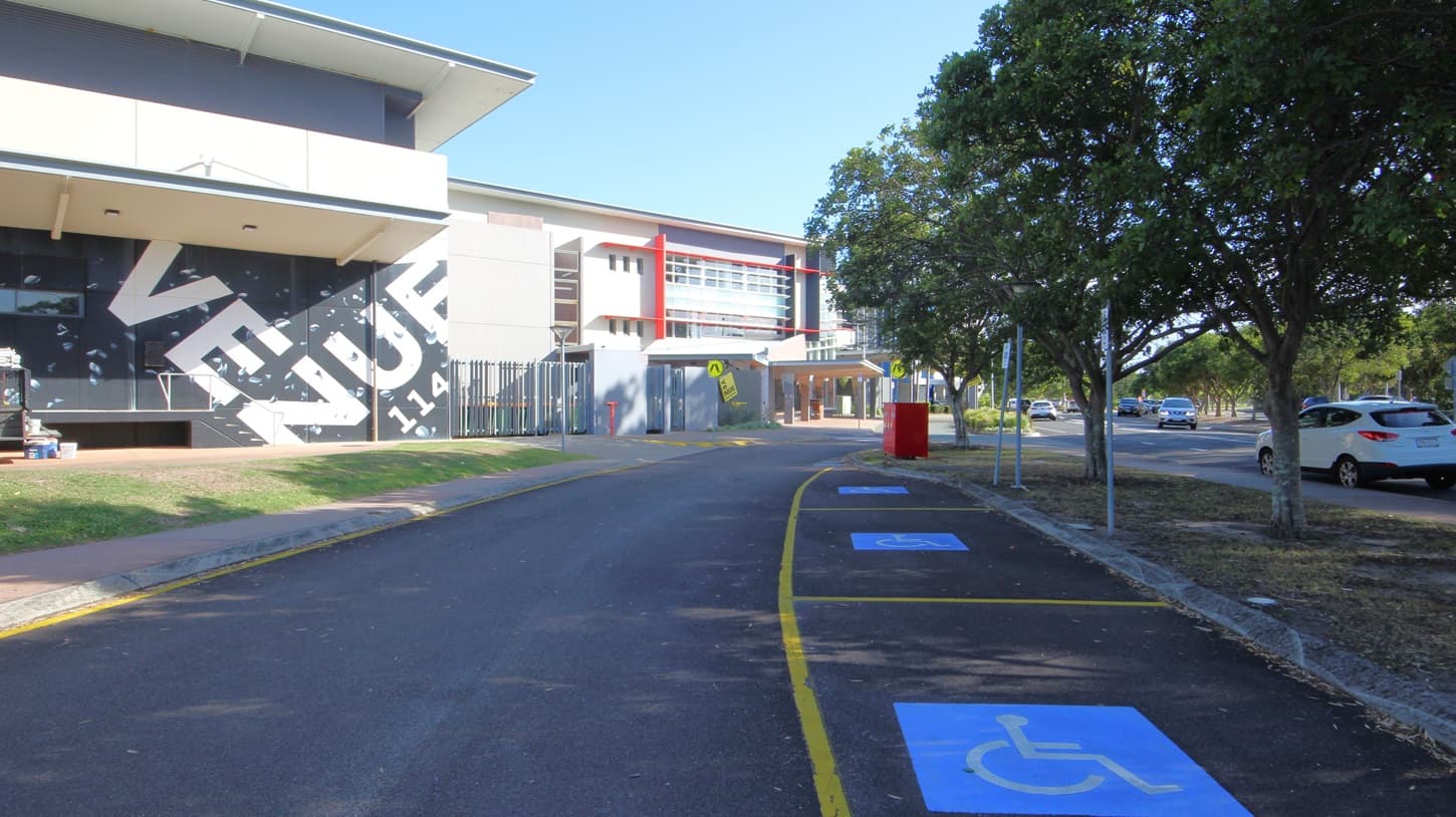 On the left, there are white pillars supporting a white ceiling with recessed lights, and glass doors leading into a building. On the right, there is an open grassy area with a palm tree. In the middle, there is a walkway with a grey concrete floor extending towards a fenced area. The background includes a building with a black fence and a view of a body of water with trees along the shoreline.