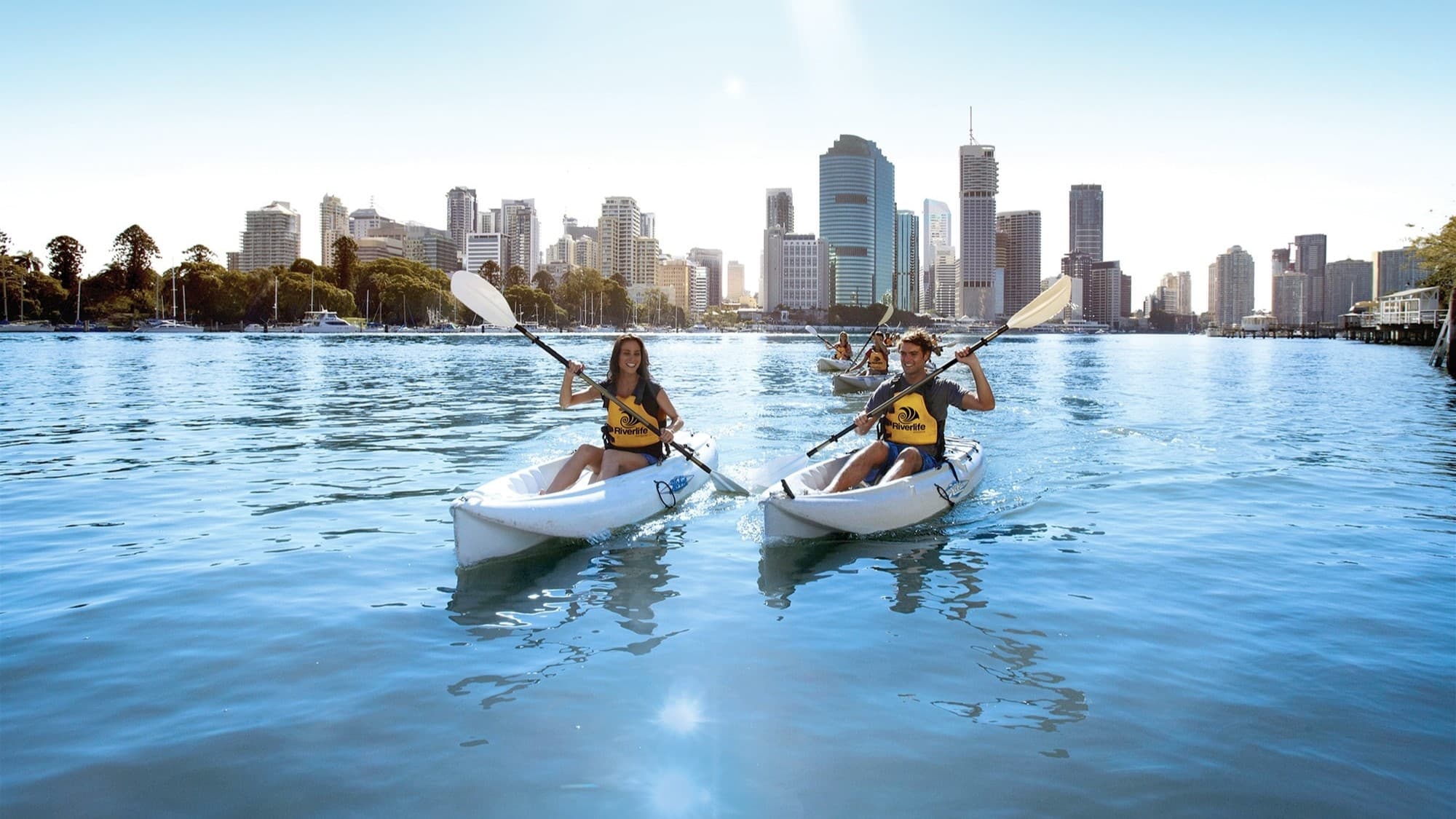 Three kayaks on the Brisbane river with the Brisbane CBD in the background.  The kayaks are white in colour and water appears a dark blue with skyline reflecting on the waters surface.