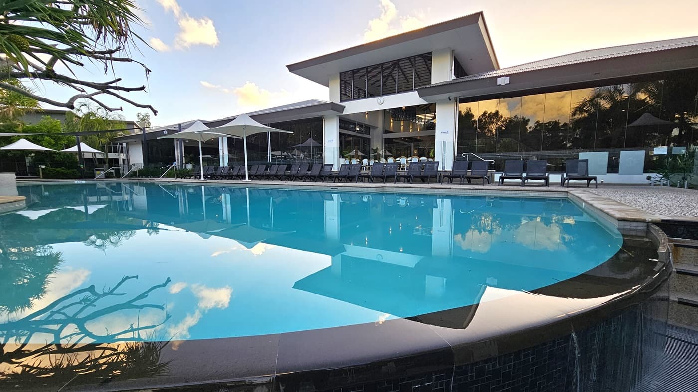 In the middle is a large pool with sun loungers lining the pool deck. A building made of concrete and glass stands behind it. Large white umbrellas cover a section of the pool. To the right is a staircase that leads to the upper section of the pool deck. A tree casts its shadow on the pool water.
