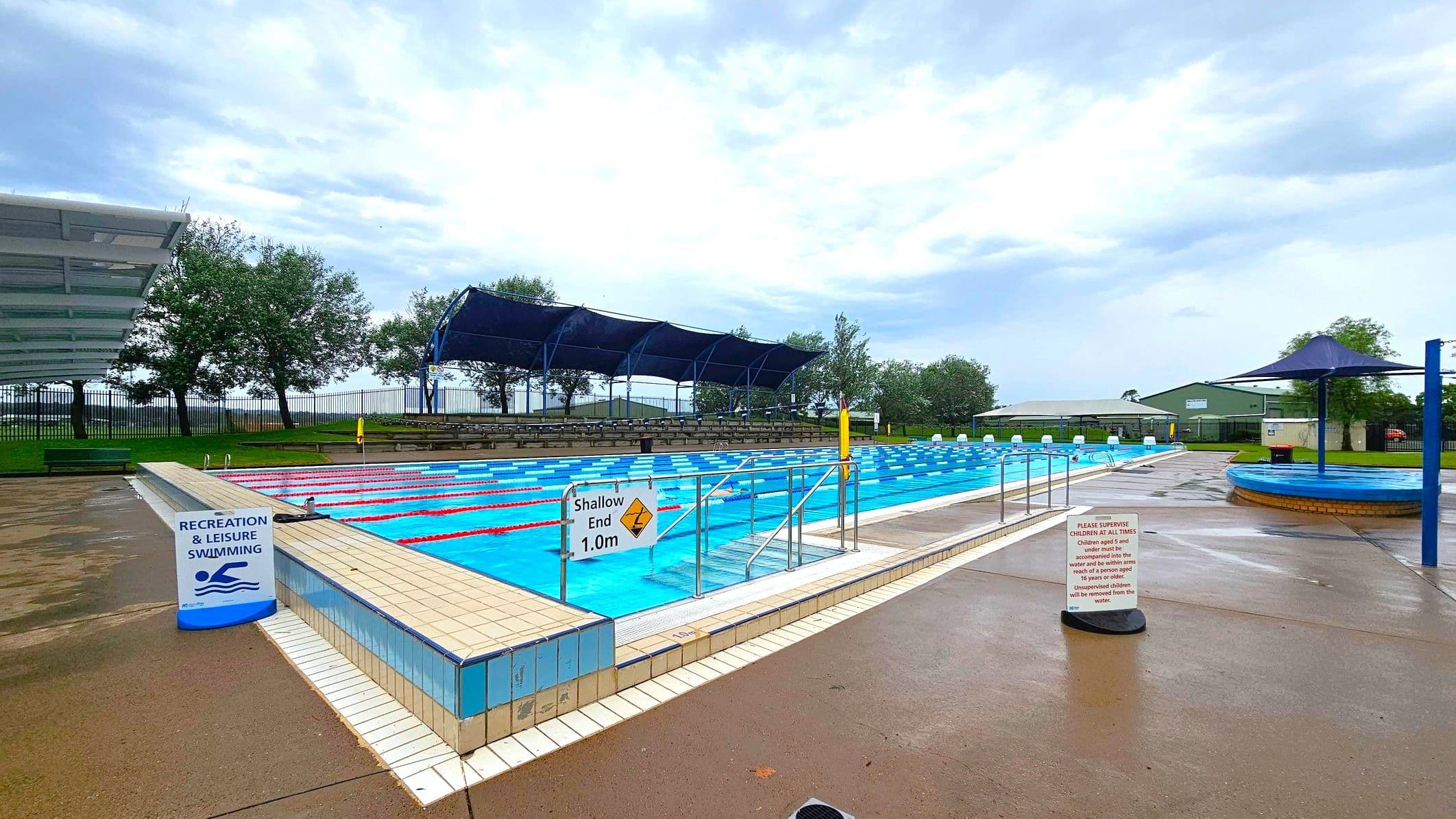 An outdoor swimming pool with multiple lanes, marked by red and blue lane dividers. On the left, there is a wet concrete walkway with a black bin and a lifebuoy. On the right, a covered area with a transparent roof and two people in red uniforms standing near signs that read "Lap Swimming." In the background, there are trees, a fence, and some buildings. The pool's edge is tiled, and a small orange cone is placed at the corner.