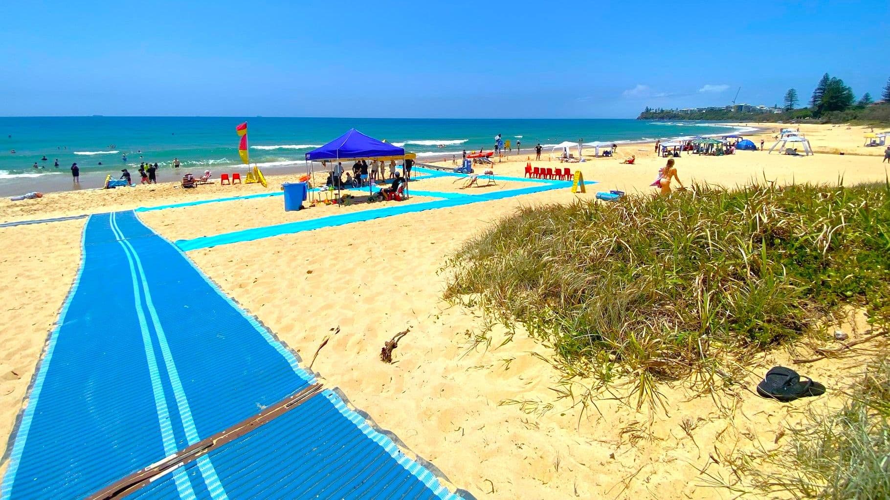 On the left, a large pandanus tree with thick foliage rests on sandy ground near a wooden post with wire fencing. On the right, a blue sign on a metal post reads "Accessible Beach Access Point" above sand and grass. In the middle, a blue mobility mat stretches over the sand towards the beach, with tents and a lifeguard hut visible in the background. The floor is sandy with some grassy patches near the edges of the mat.
