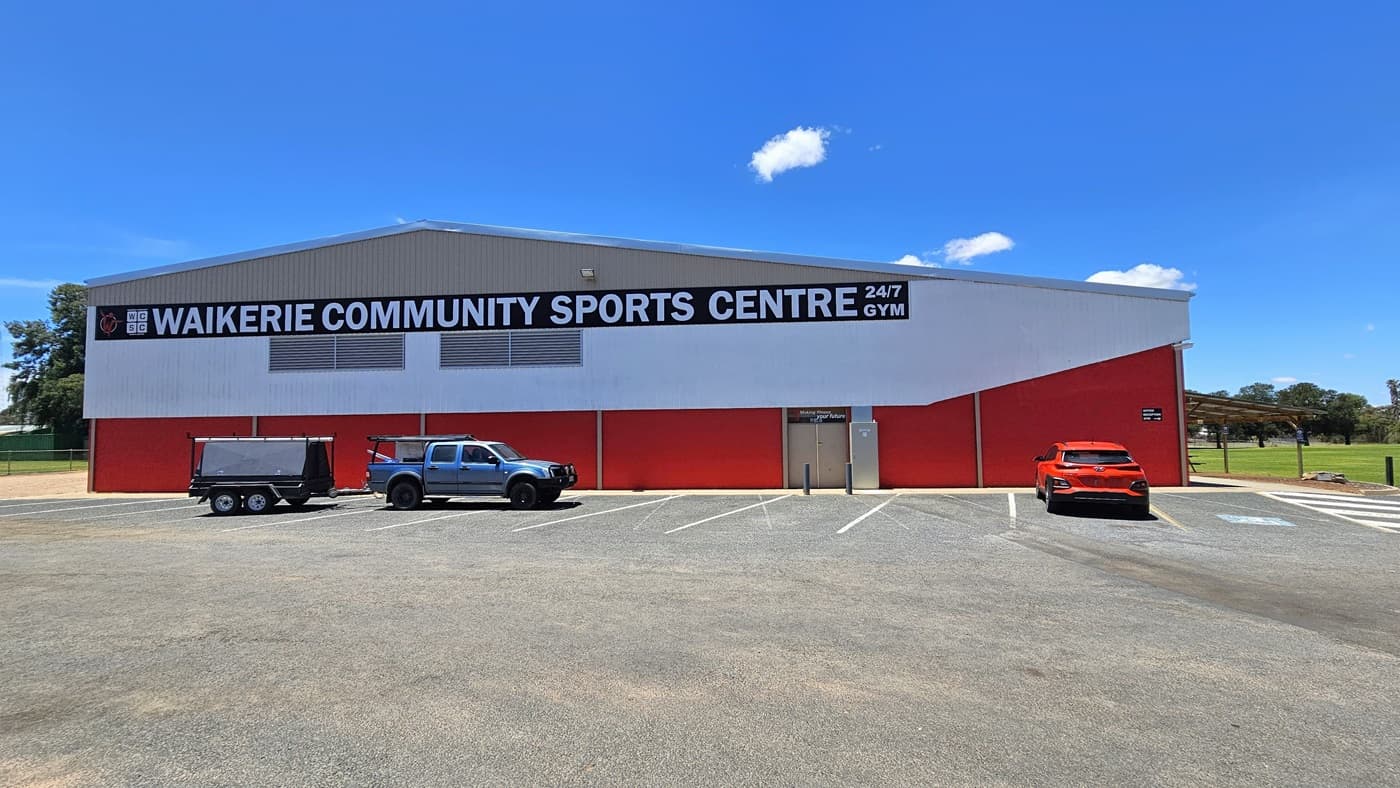 A building with a red corrugated iron and brown brick facade. Bold white letters form the prominent facility signage. The adjacent concrete car park accommodates three cars of various models and colours, with designated accessible parking on the right, marked by blue wheelchairs and white diagonal lines. To the right of the building is a shaded area and an expanse of green grass.