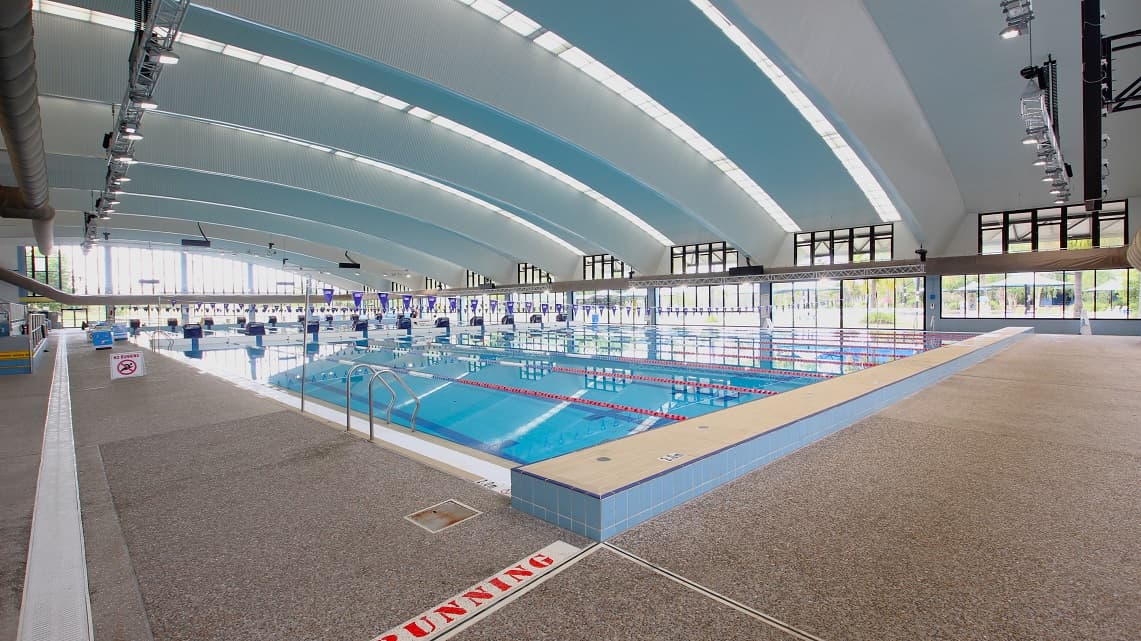 The view of the whole 50m pool from the benches. Beneath the benches, you can observe metallic fencing surrounding the pool. The expansive 50-meter pool is distinctly visible from the seating area. The indoor pool appears to undergo a regular and thorough cleaning. Additionally, blue pool flags are extended across the pool from the opposite side.
