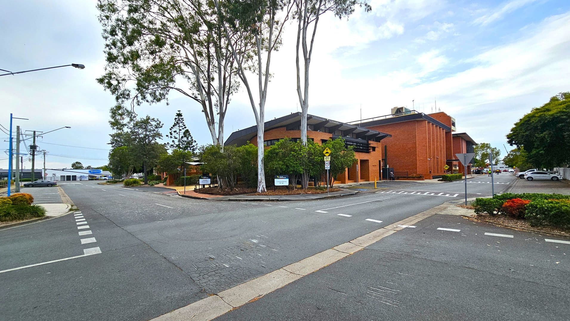 A street scene with a brick building on the left, featuring a covered entrance and surrounded by trees and shrubs. A large tree stands near the building. On the right, there is a parking area with marked spaces, including a designated disabled parking spot. The background shows more trees, a fence, and a few buildings. A streetlight and utility pole are visible near the parking area. The road is empty, and the pavement is bordered by a narrow footpath.