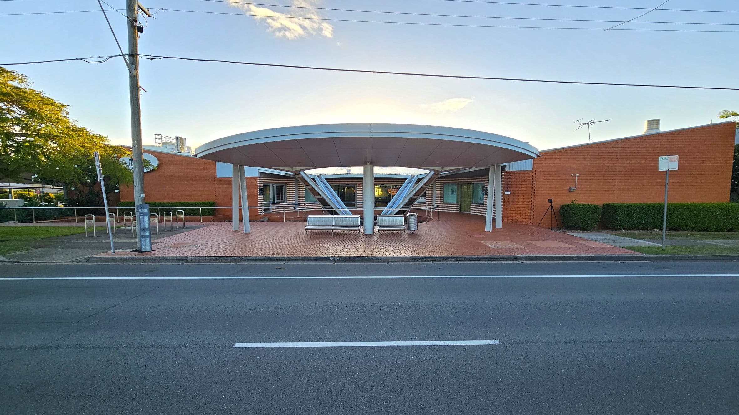 The image shows the entrance to a building with large glass windows and a colourful mural on the wall to the right. A bench is positioned near the entrance, and a pathway with a railing leads up to the door. The building is surrounded by greenery, with trees visible in the background. The ceiling above the entrance is grey, and the ground is paved with concrete.