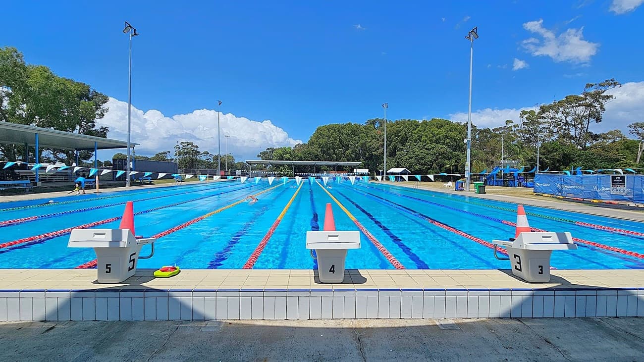 An outdoor swimming pool with multiple lanes, each marked by red and yellow lane dividers. A diving block is visible in the foreground. On the left, there are white benches and a building with a blue umbrella. On the right, trees and a shaded seating area are present. Tall floodlights are positioned around the pool, and a row of triangular flags stretches across the lanes. The pool area is surrounded by grass.