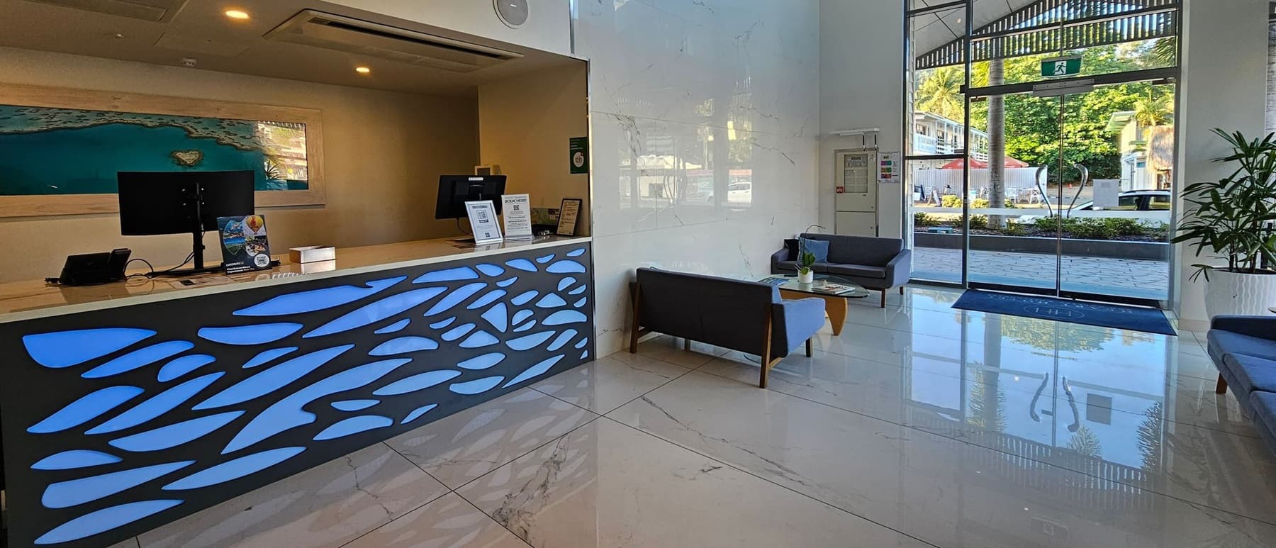 A modern reception area with a sleek desk on the left, featuring a blue abstract design and two computer monitors. Behind the desk is a large framed image of a coastal scene. On the right, there are glass doors leading outside, with a view of greenery and buildings. Two grey sofas and a wooden coffee table are positioned near the entrance. The floor is polished marble, and the ceiling is high with recessed lighting.