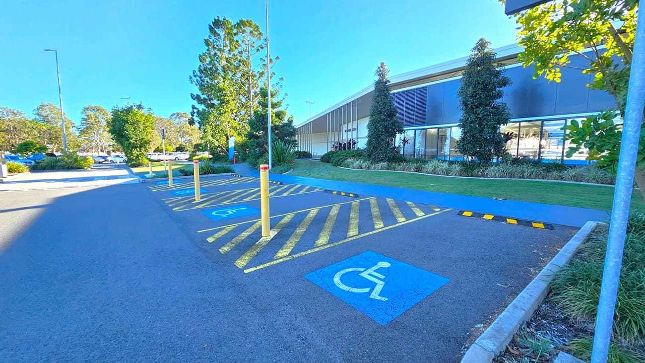 Accessible parking spaces with blue wheelchair symbols on the ground are on the right. Yellow-striped zones separate the spaces, with yellow bollards in front. A modern building with large windows and landscaped greenery is in the background. Trees and shrubs line the area, and a few cars are visible in the distance on the left. The sky is clear and blue.